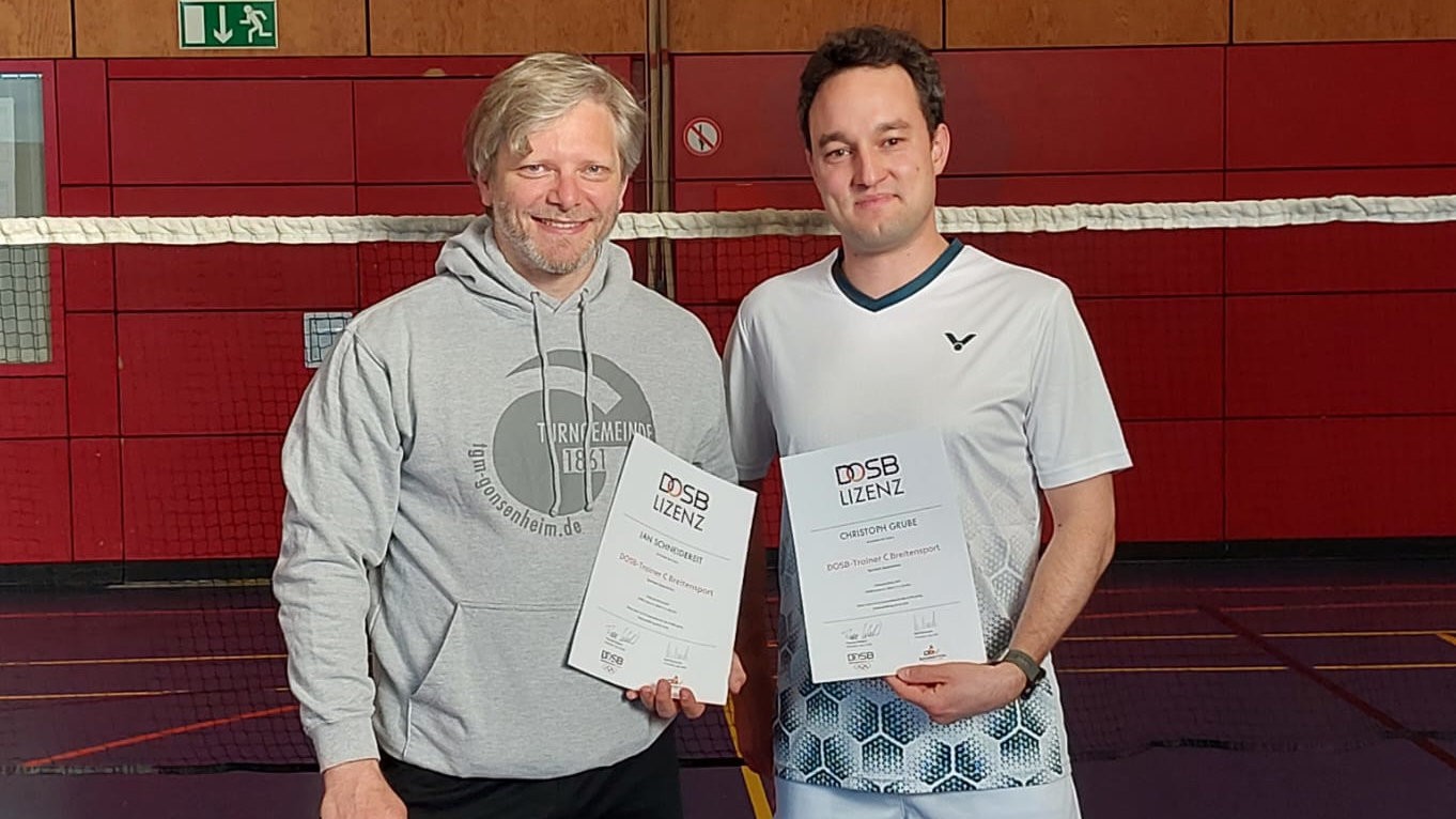 Zwei Männer halten Zertifikate nach bestandener Trainerausbildung in einer Sporthalle.
