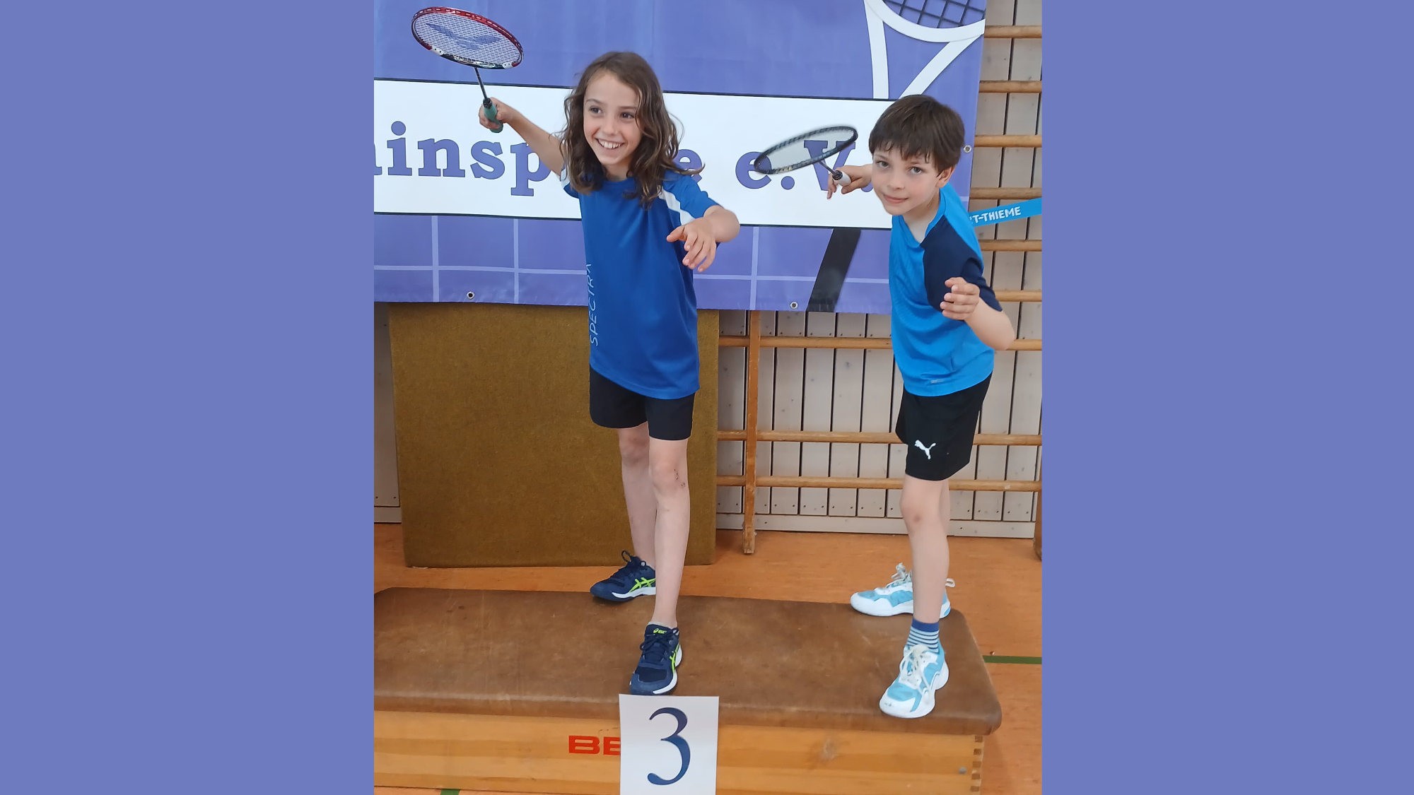 Zwei Kinder in blauer Sportkleidung feiern auf einem Podest bei einem Badminton-Turnier.