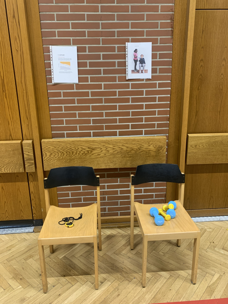 Vorbereitung für AFT-Test: Stühle mit Hanteln und Stoppuhr in der Turnhalle Zwei Stühle mit Fitnessgeräten wie Hanteln und Stoppuhr vor einer Turnhallenwand, vermutlich für einen AFT-Test oder Fitness-Test vorbereitet.