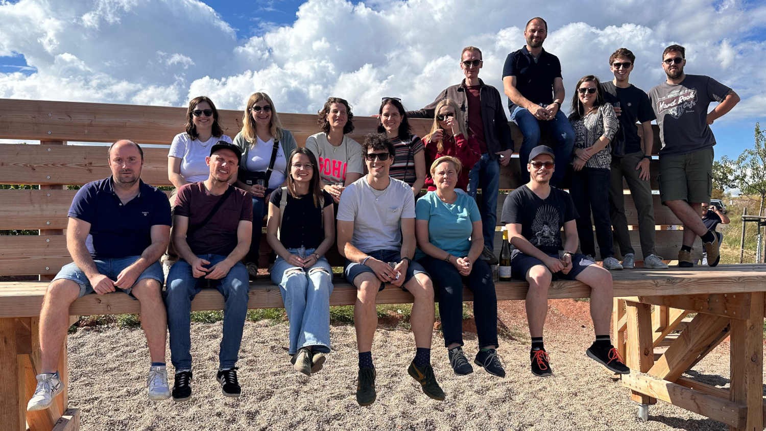 Gruppe von Menschen bei einer Sommeraktivität im Freien auf einer Holzbank unter blauem Himmel