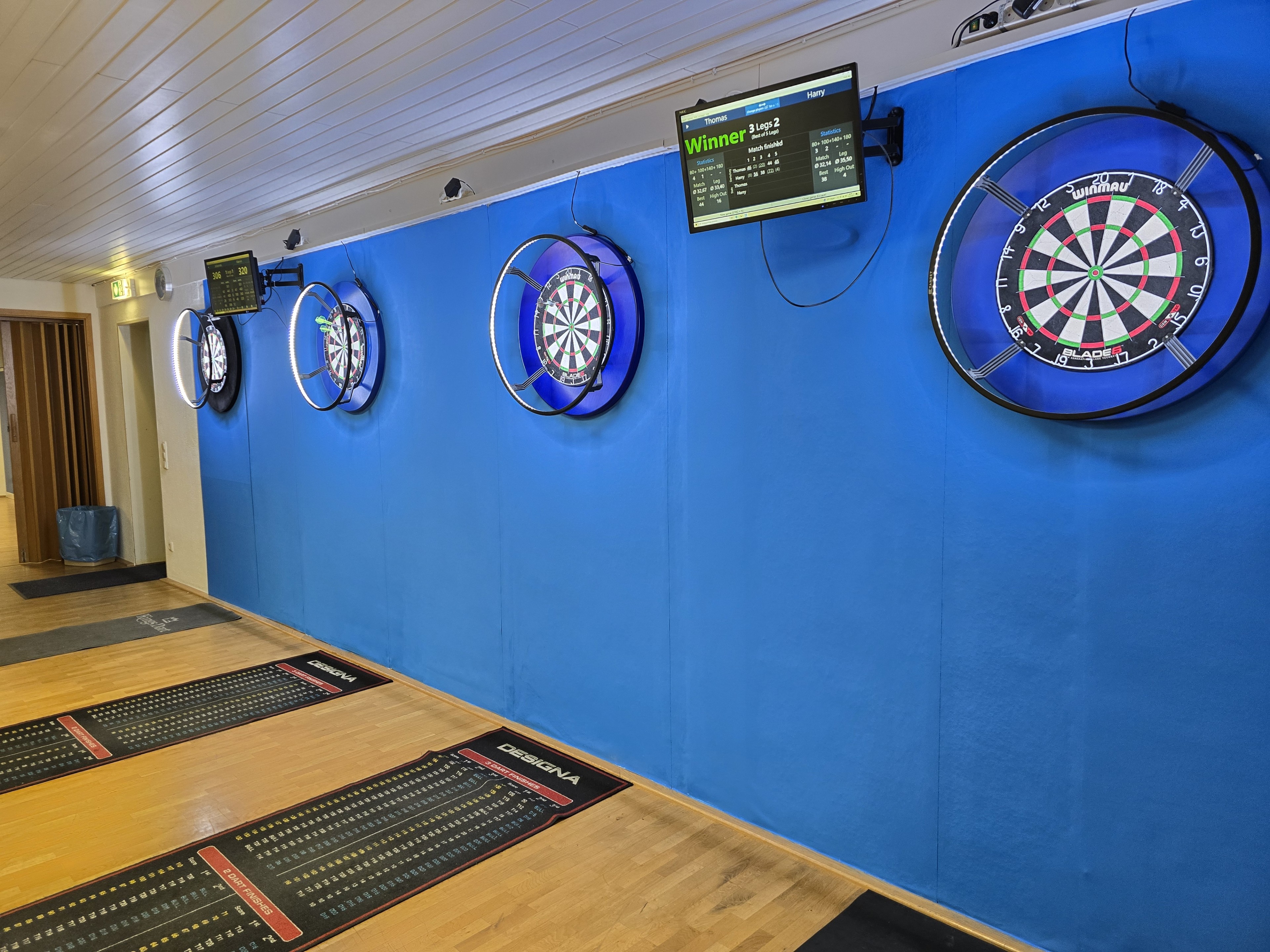 Dartboards und elektronische Anzeigetafeln in einem modernen Darts-Trainingsraum mit blauer Wand und Designa-Matten.