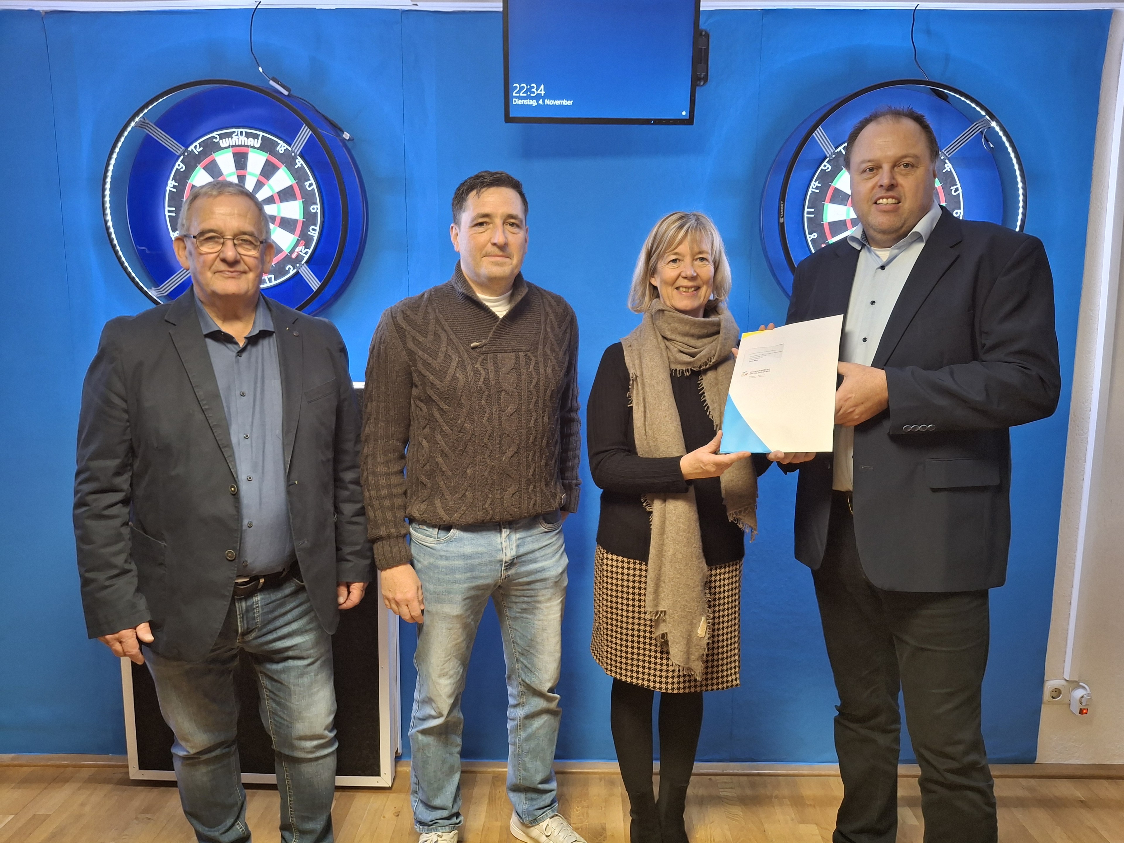 Vier Personen stehen in einem Raum mit blauer Wand, zwei Dartscheiben und einem Bildschirm, eine Person überreicht ein Dokument.