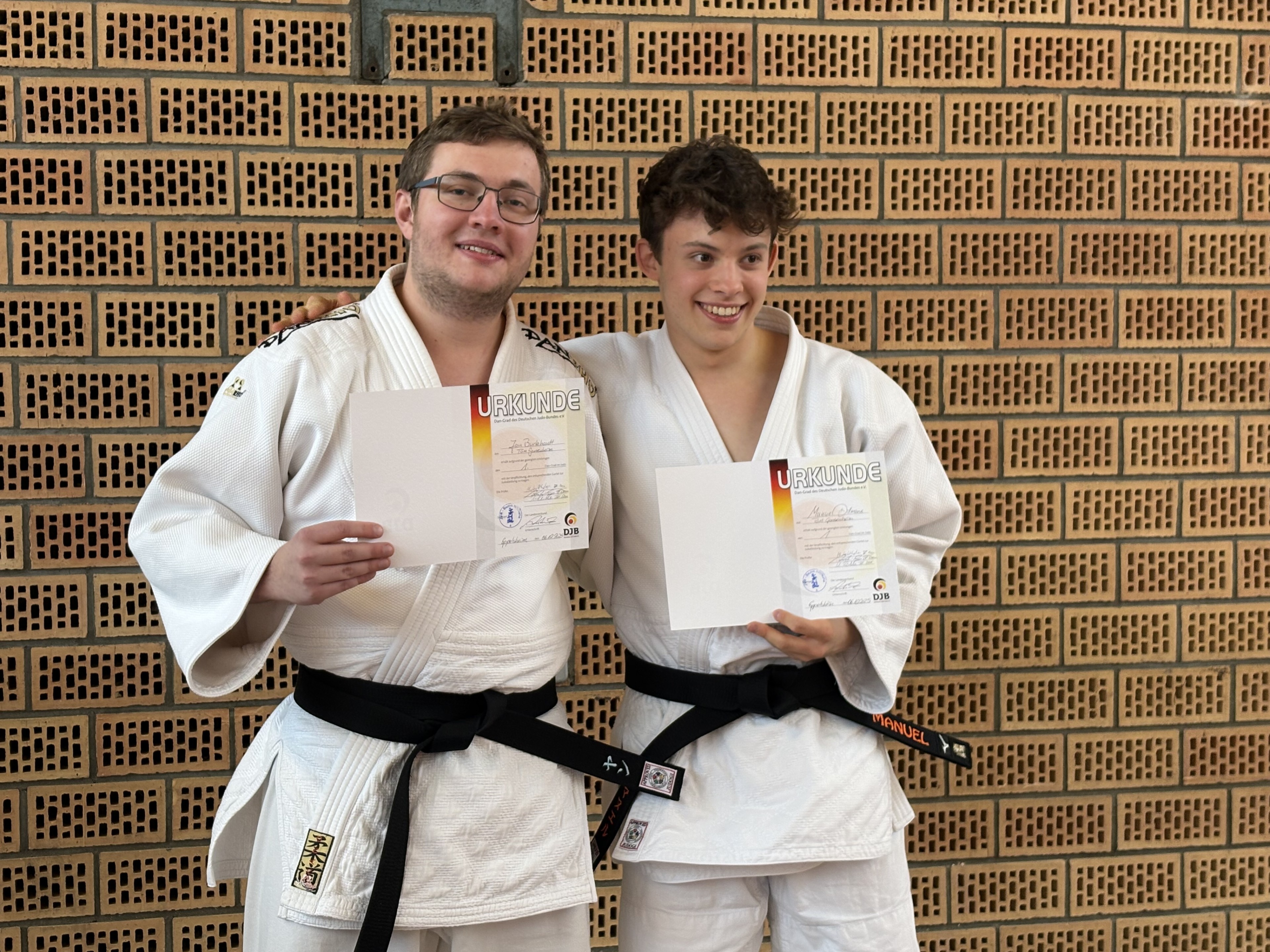 Zwei Judoka mit schwarzem Gürtel halten Urkunden vor einer Backsteinwand.