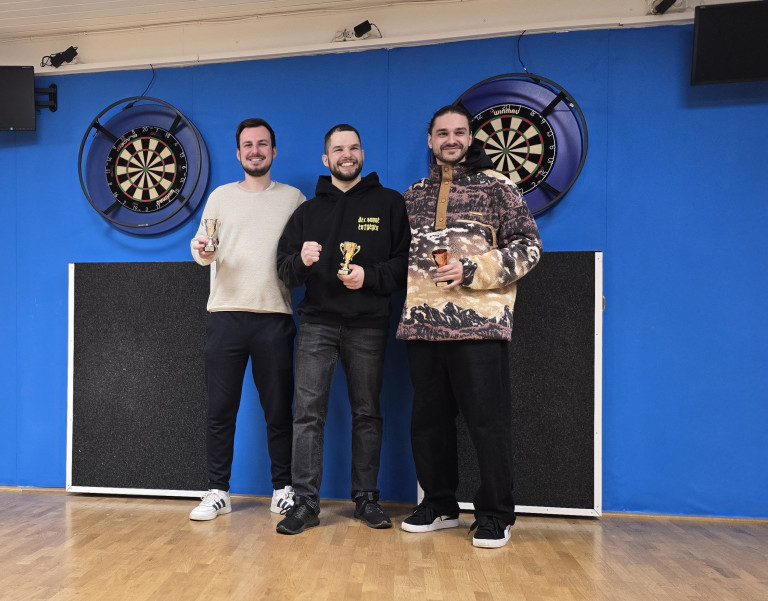 Siegerehrung 2025 – Gewinner mit Pokalen vor Dartboards Siegerehrung 2025 mit drei Gewinnern vor Dartboards und Pokalen