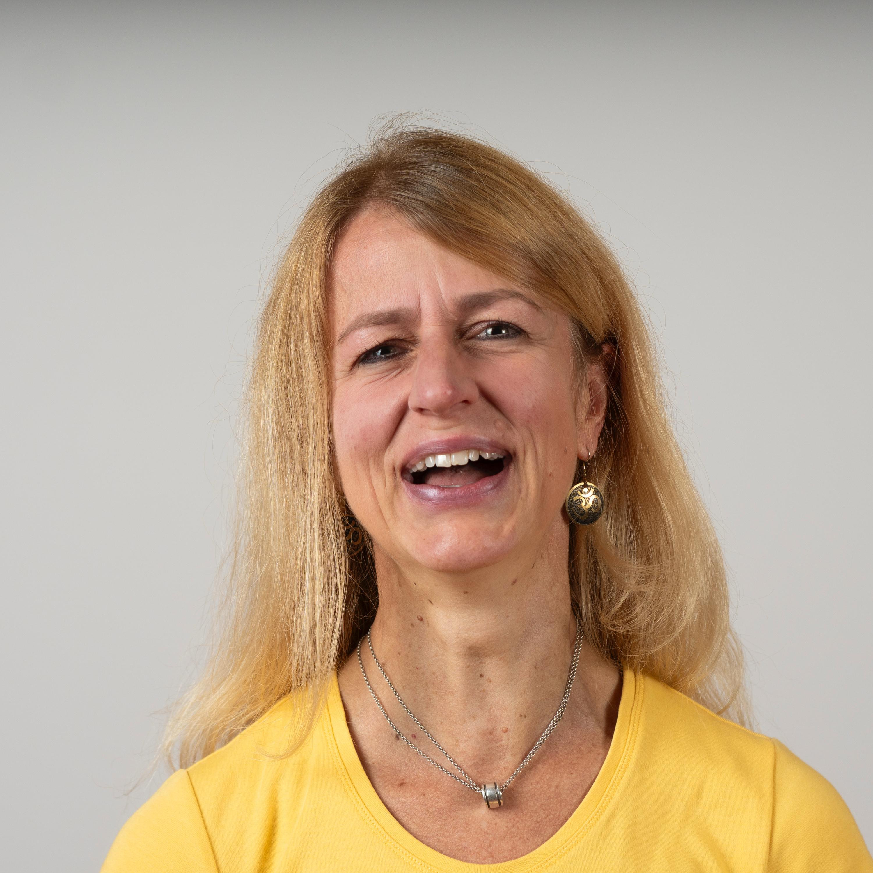 Frau mit langen blonden Haaren und gelbem T-Shirt, trägt Schmuck vor neutralem Hintergrund