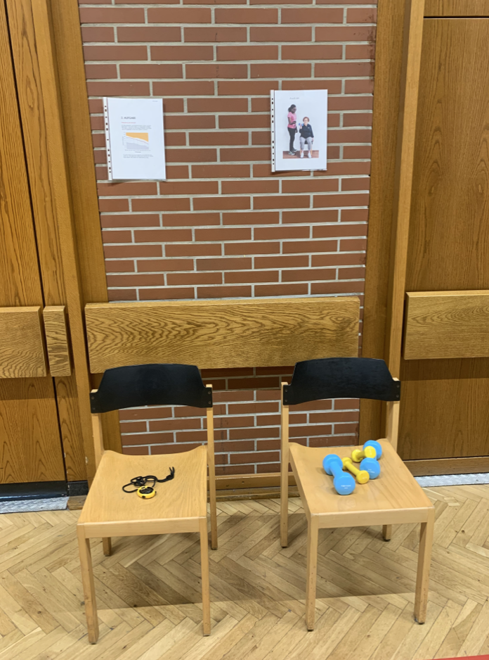 Zwei Stühle mit Fitness-Equipment wie Hanteln und Springseil vor einer Wand, möglicherweise für ein AFT-Training oder eine Fitnessübung.