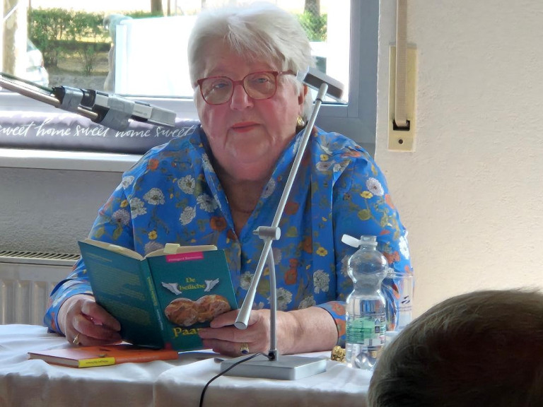 Ältere Frau liest bei einer Lesung aus einem Buch mit dem Titel 'Die besten Paare' vor, Mikrofon und Wasserflasche auf dem Tisch