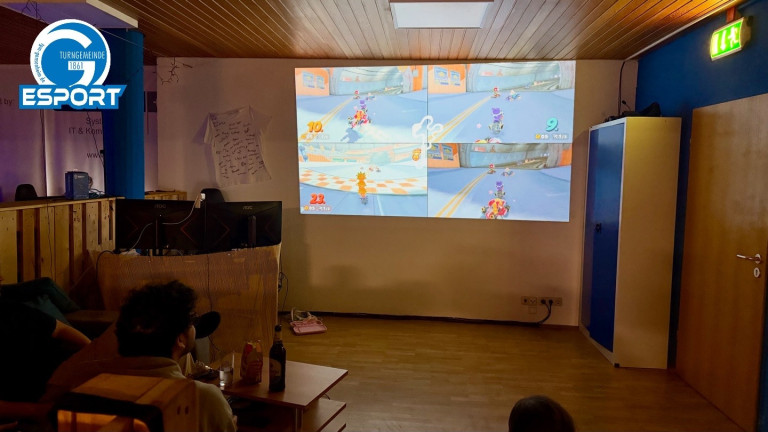 Mehrere Personen spielen ein Videospiel im Splitscreen-Modus auf einer großen Leinwand in einem Wohnzimmer.