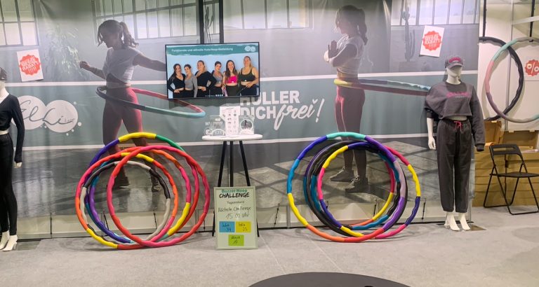 Messestand mit Hula-Hoop-Reifen, Werbeplakat und digitaler Anzeige, Buzzer-Hoop Challenge Tagesrekord und nächste Challenge um 15 Uhr.