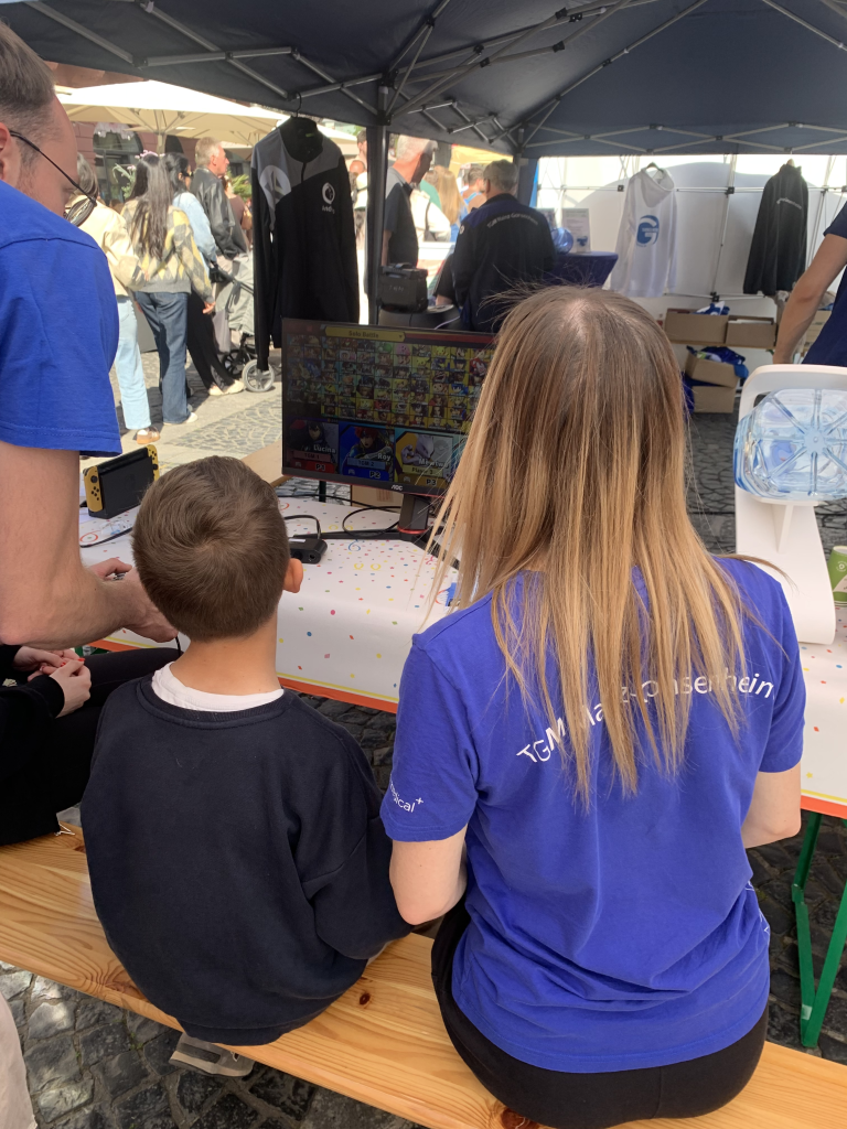 Teilnehmer spielen Videospiel auf der E-Sports Meile des Sports an einem Stand mit mehreren Personen im Hintergrund.