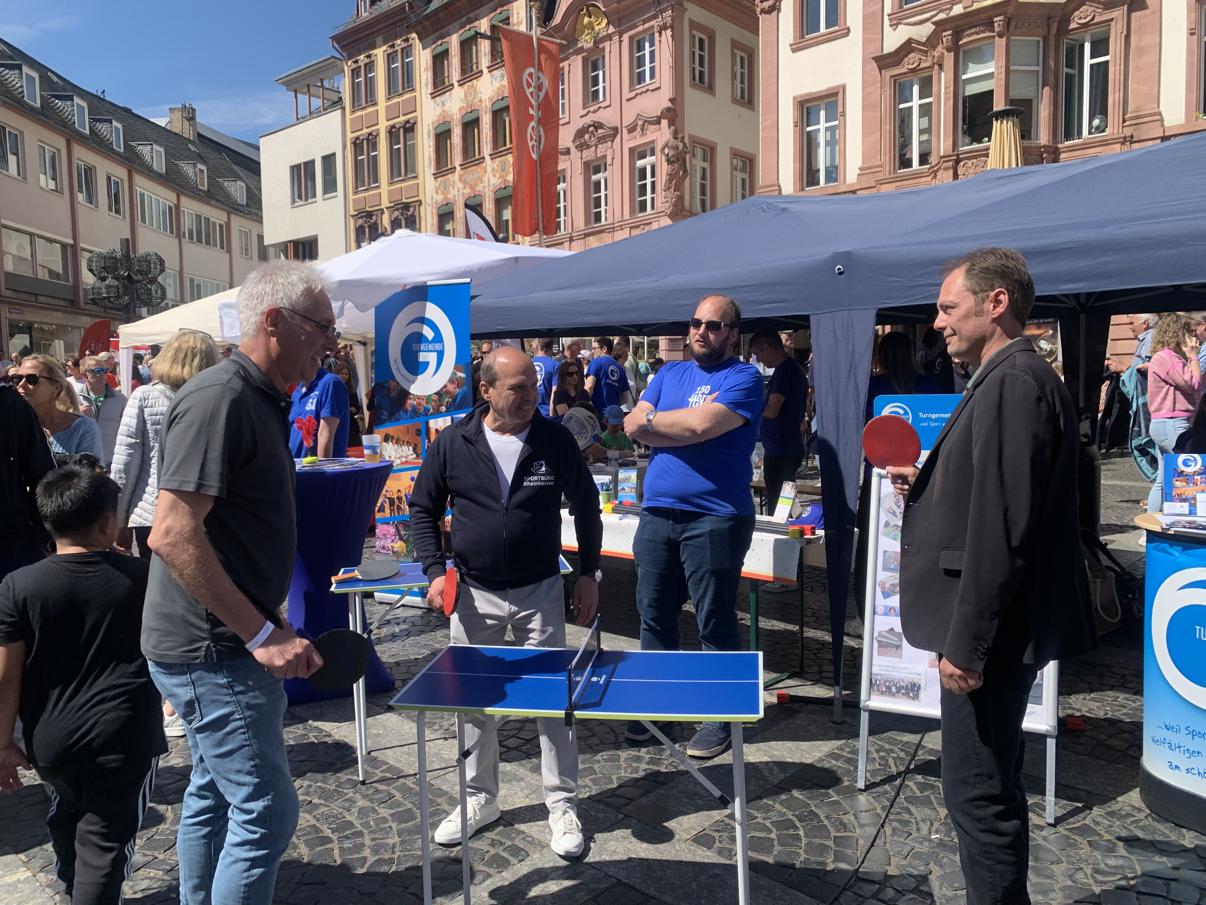 Stadtsportverband Meile des Sports: Menschen spielen Tischtennis auf einem Stadtfest mit Infoständen im Hintergrund