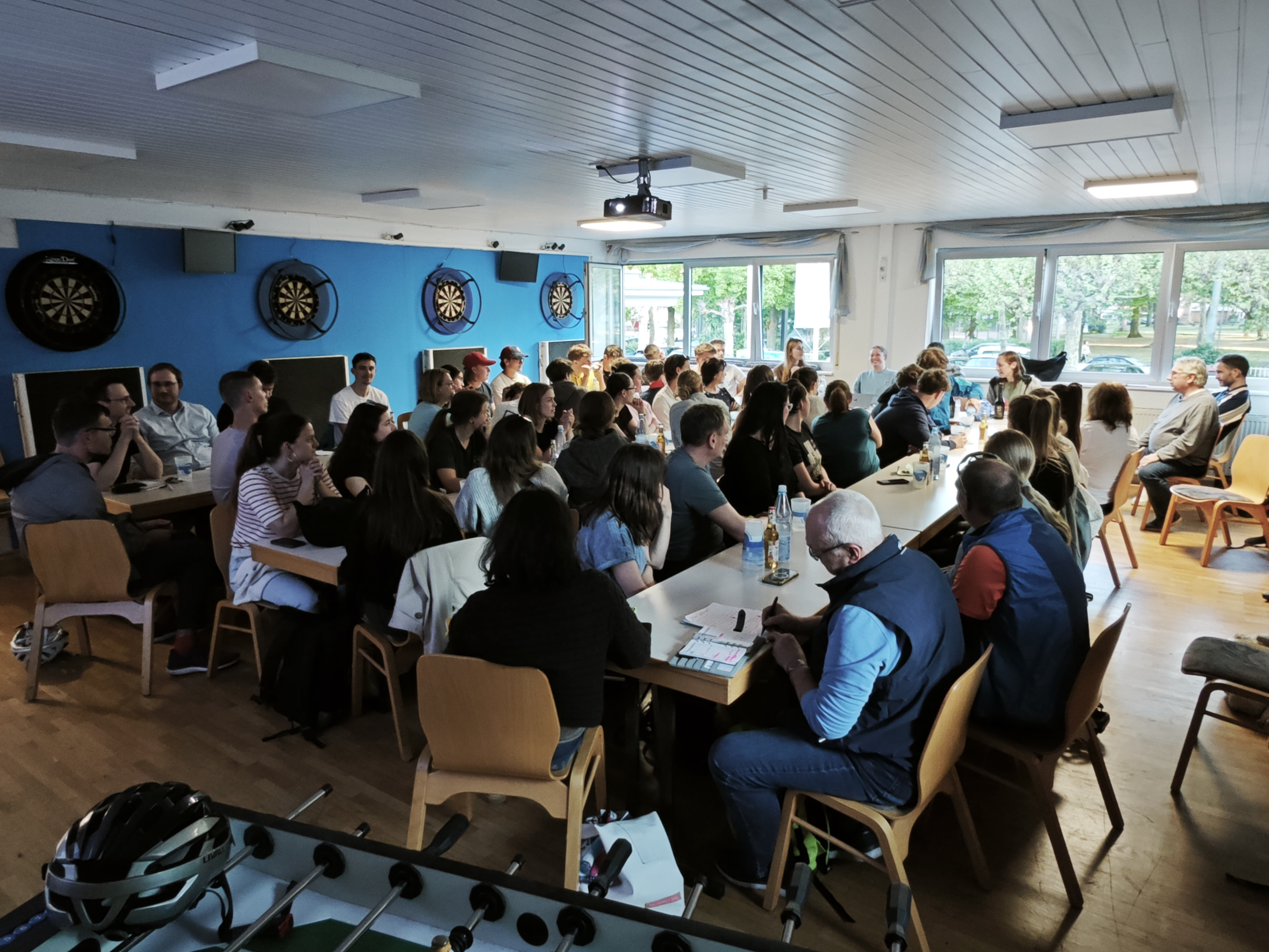Große Gruppe von Menschen bei einer Veranstaltung in einem Gemeinschaftsraum mit Dartscheiben an der Wand, aufgenommen am 23.04.2026