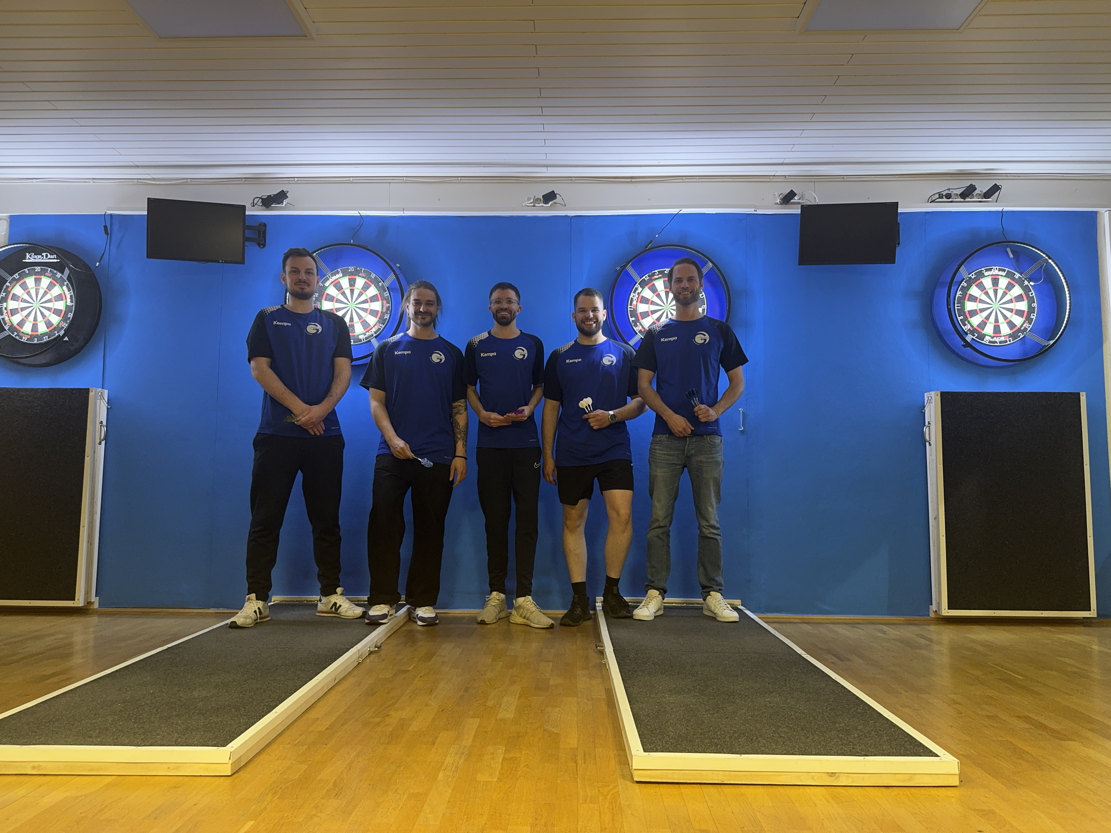 Fünf junge Männer im blauen Trikot stehen vor Dartscheiben in einem Dartverein