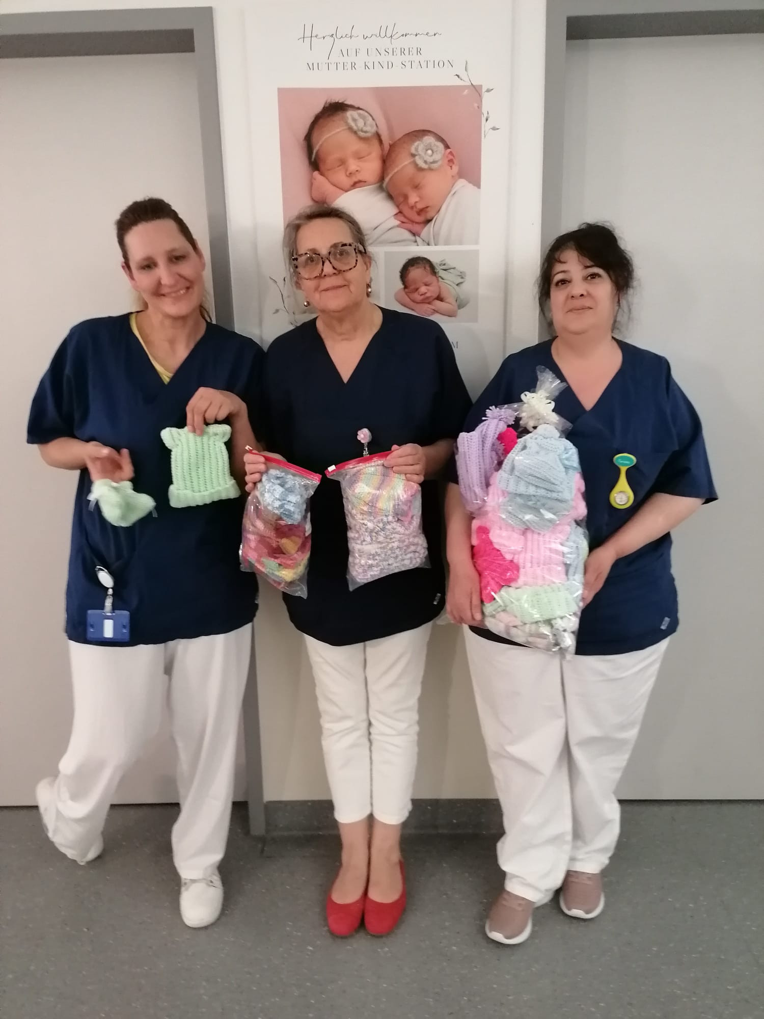 Drei Pflegekräfte in blauer Dienstkleidung halten handgestrickte Babymützen und Säckchen in einem Krankenhausflur.