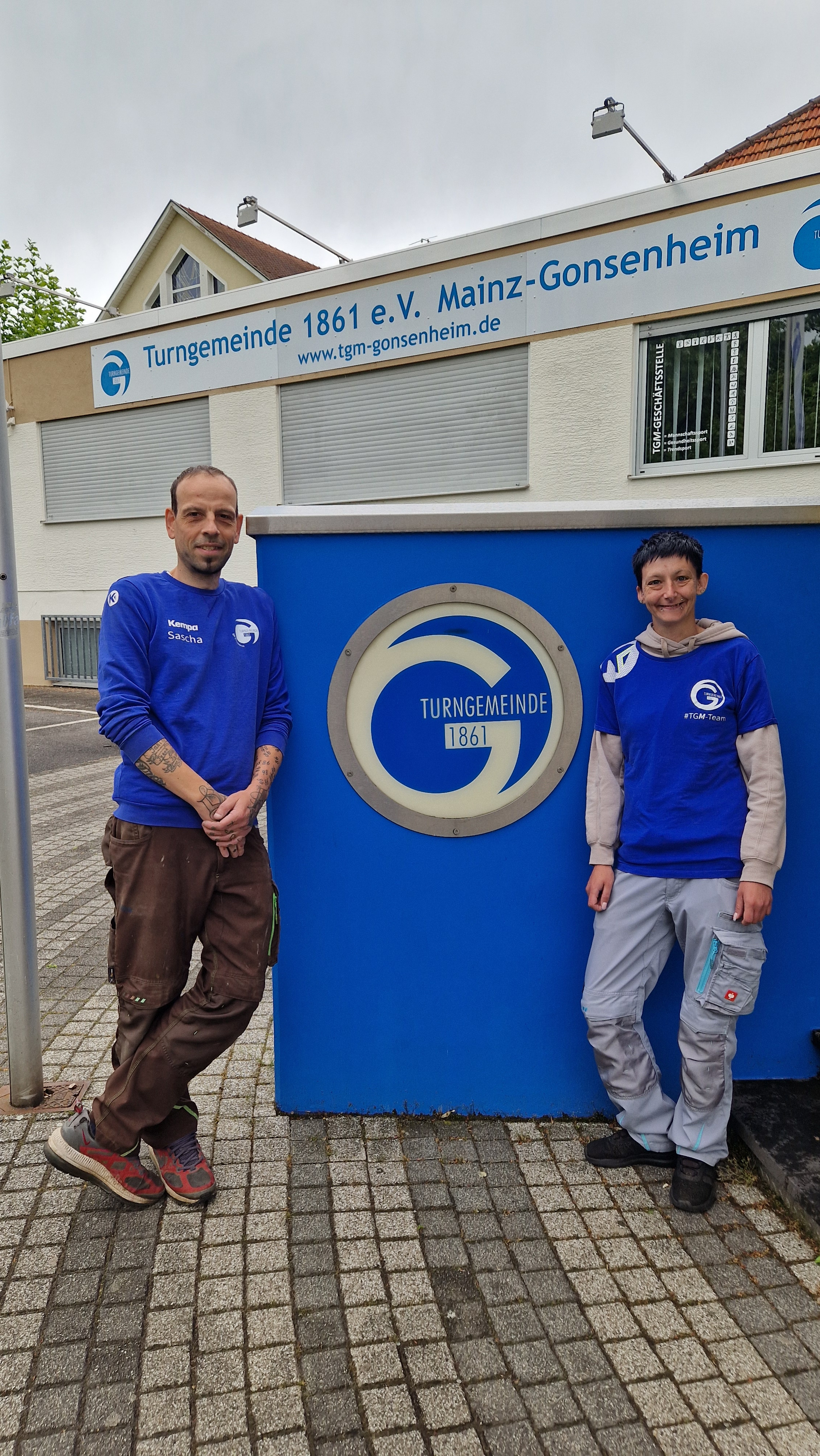 Zwei Personen vor dem Eingang der Turngemeinde 1861 Gonsenheim e.V. mit blauem Schild und Logo