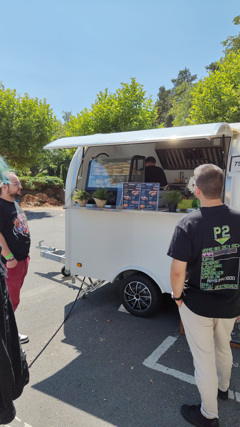 Zwei Personen stehen an einem weißen Foodtruck mit Speisekarten im Freien bei sonnigem Wetter.