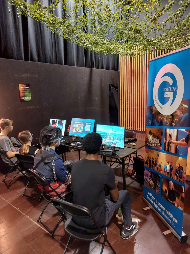 Videospiel-Event der Turngemeinde 1861 mit Jugendlichen an mehreren Monitoren Jugendliche spielen Videospiele bei einem Event der Turngemeinde 1861, mehrere Monitore und ein Banner der Turngemeinde 1861 sind sichtbar.