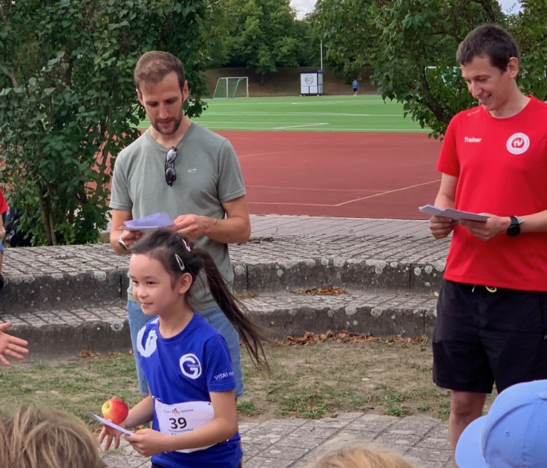 Siegerehrung bei Sportveranstaltung: Mädchen mit Urkunde und Apfel vor Trainern Mädchen erhält Urkunde bei Sportveranstaltung im Freien mit Trainern im Hintergrund