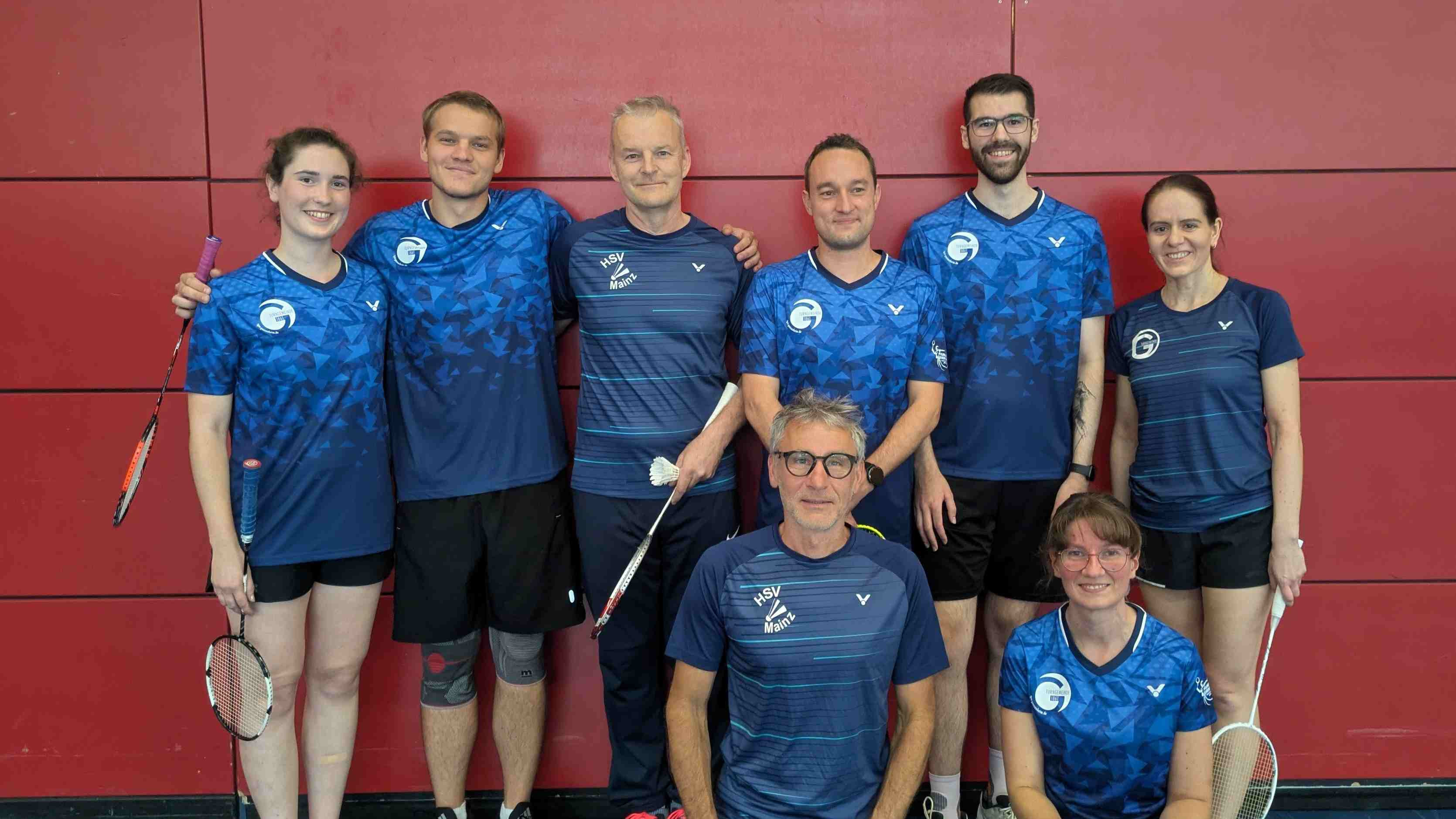 Badminton-Team II Gruppenfoto vor roter Wand, Spieler in blauen Trikots