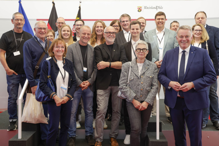 Gruppenfoto in der Staatskanzlei Rheinland-Pfalz, aufgenommen beim Sommer-Event, mehrere Personen vor dem Rheinland-Pfalz-Logo