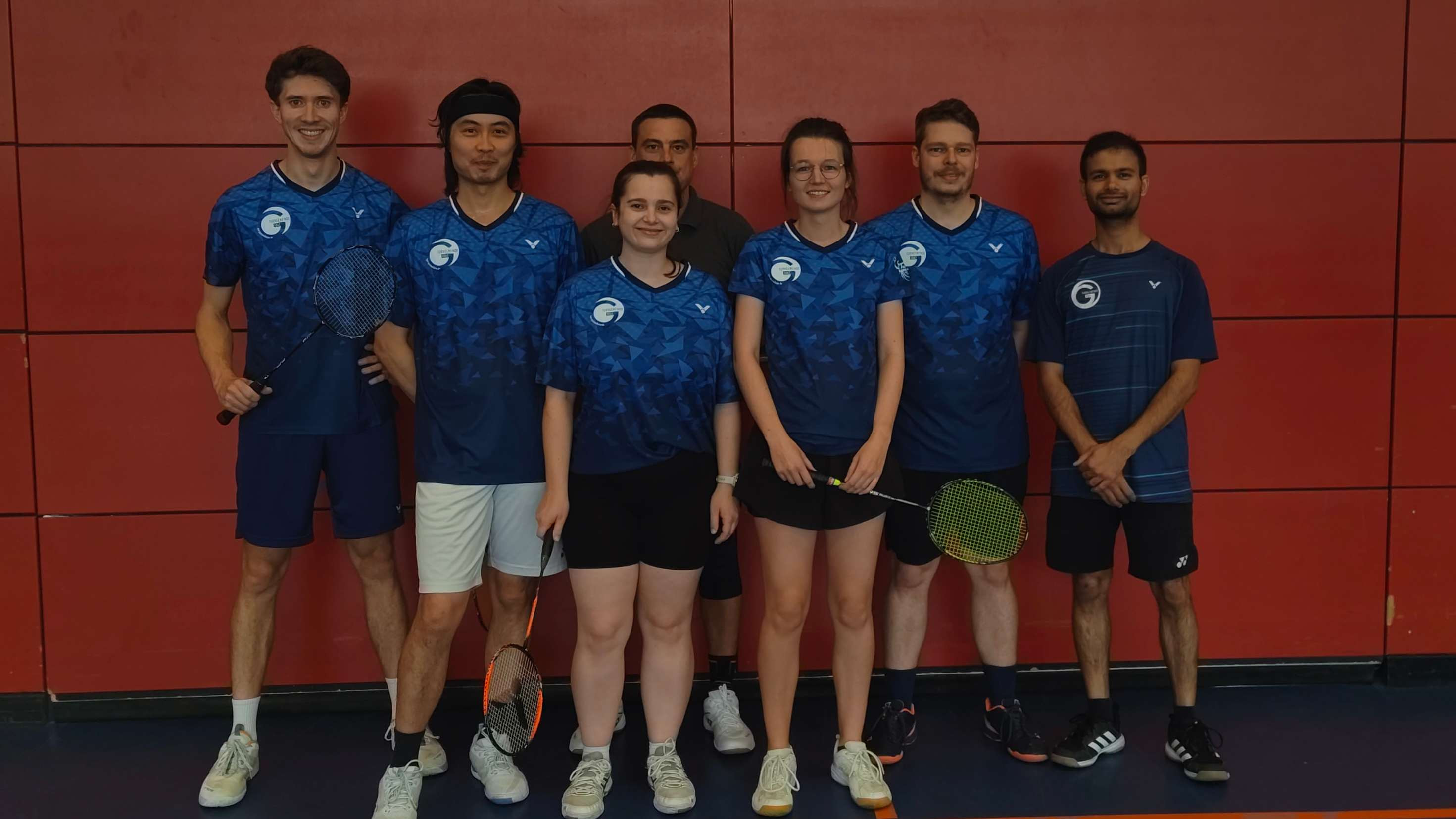 Gruppenfoto der 3. Mannschaft eines Badminton-Teams in blauen Trikots vor roter Wand