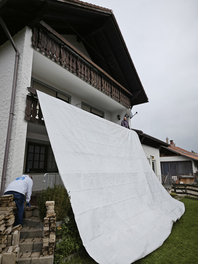 Große weiße Plane wird von zwei Personen an einem Balkon eines Hauses angebracht Zwei Personen befestigen eine große weiße Plane an einem Balkon eines Einfamilienhauses