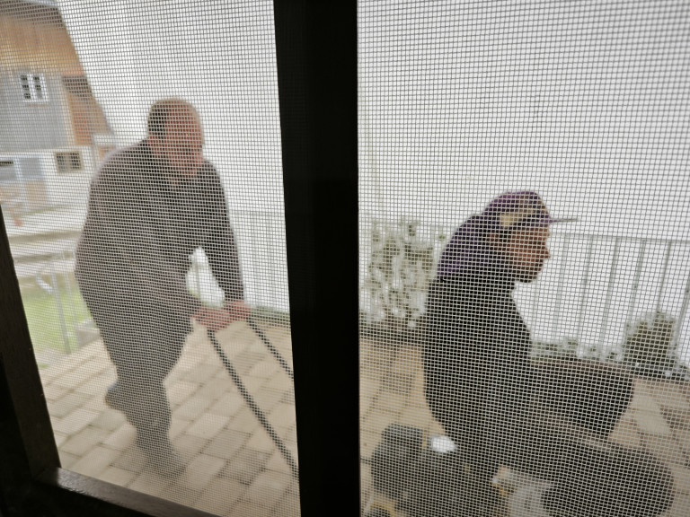 Zwei Männer bei der Arbeit auf einer Terrasse, Blick durch ein Fliegengitter Zwei Männer arbeiten draußen auf einer Terrasse, fotografiert durch ein Fliegengitter
