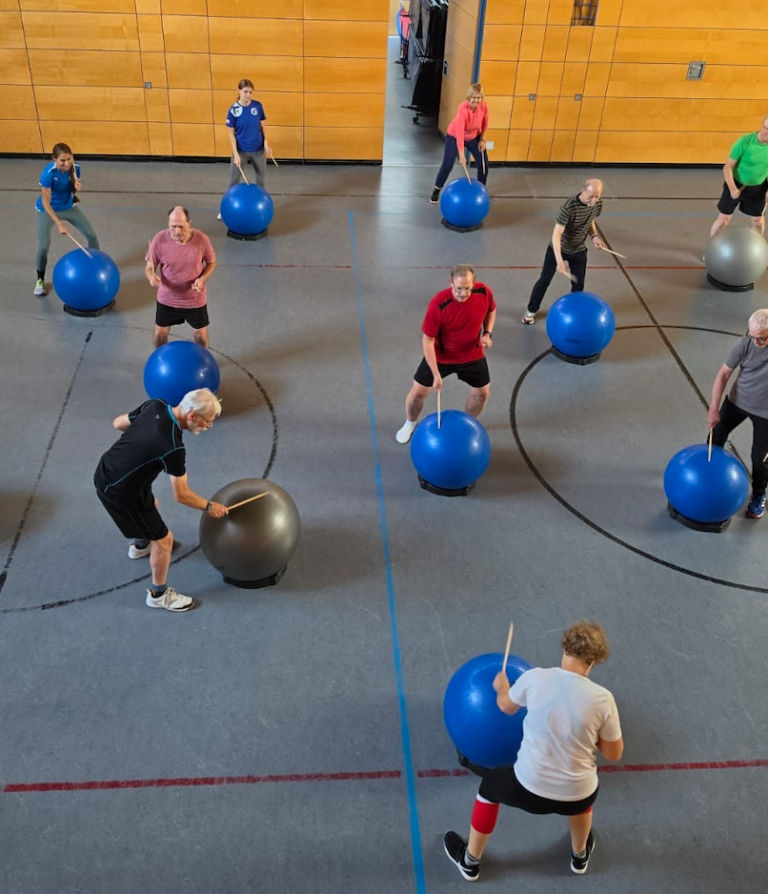 Senioren nehmen an einem Drums Alive Herzsport-Kurs in einer Sporthalle teil und trommeln auf Gymnastikbällen.