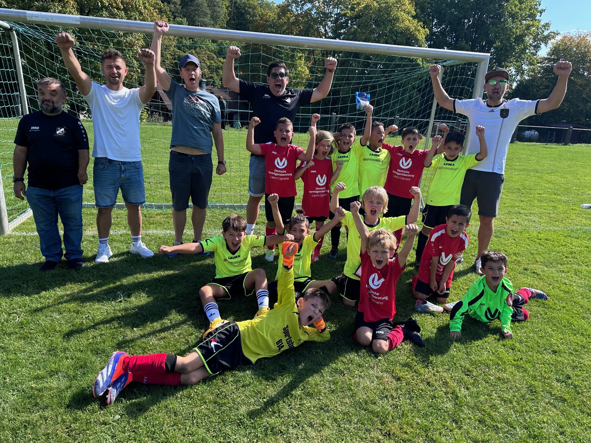 Kinder-Fußballmannschaft mit Trainern jubelt auf dem Sportplatz vor dem Tor