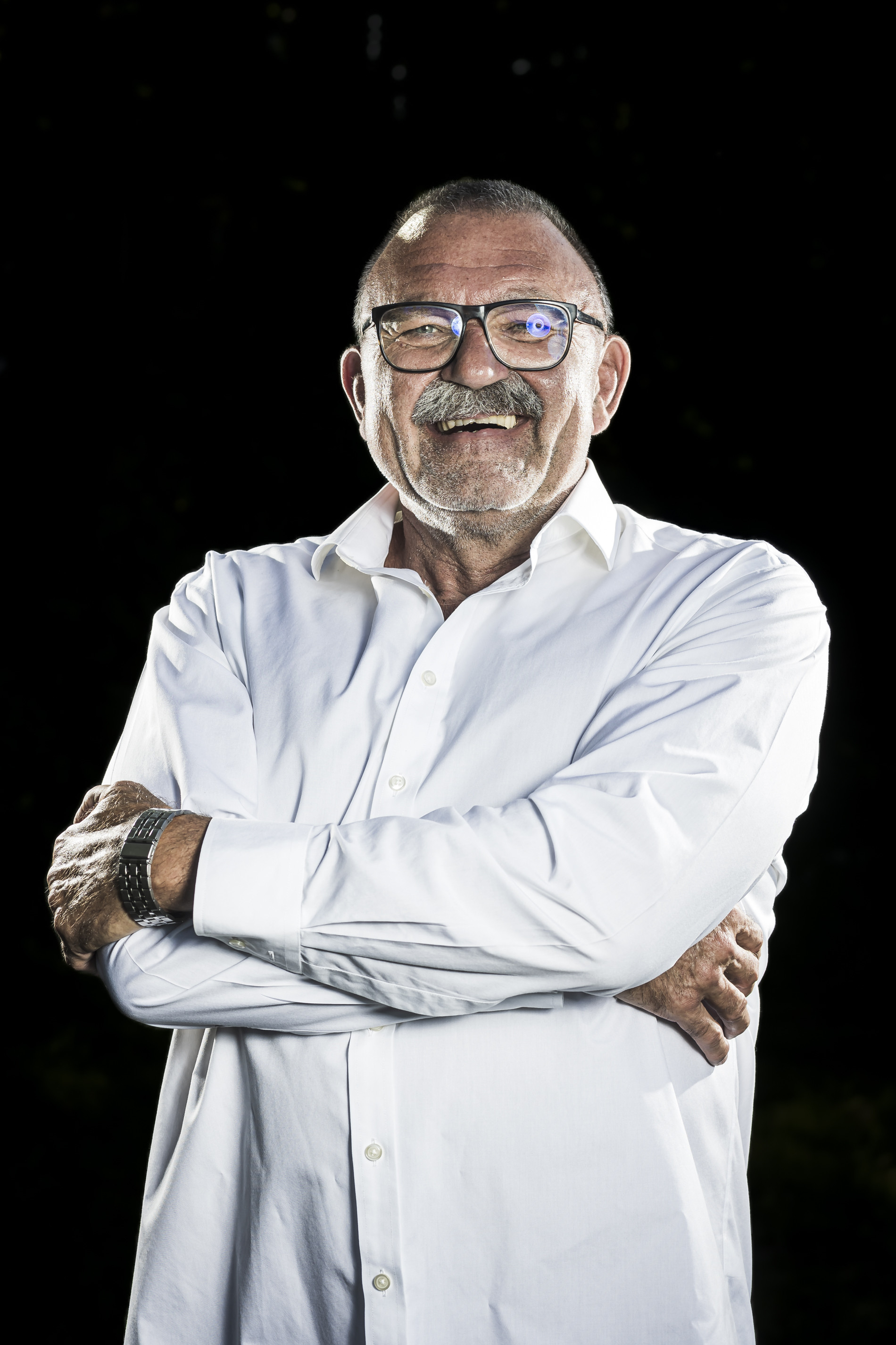 Älterer Mann mit Brille und weißem Hemd lächelt mit verschränkten Armen vor schwarzem Hintergrund