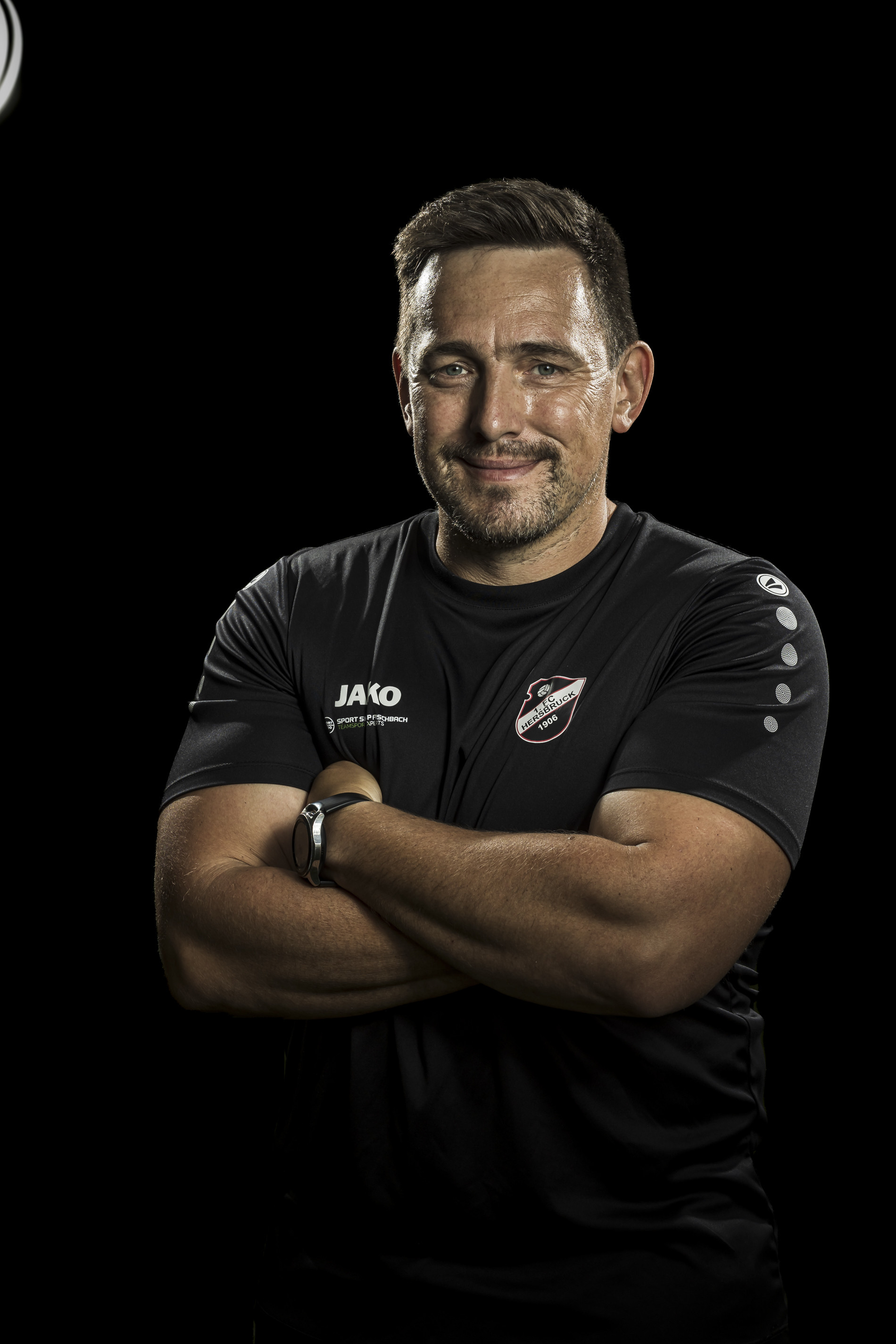 Porträt eines Mannes im schwarzen Sportshirt mit Vereinslogo und verschränkten Armen vor schwarzem Hintergrund