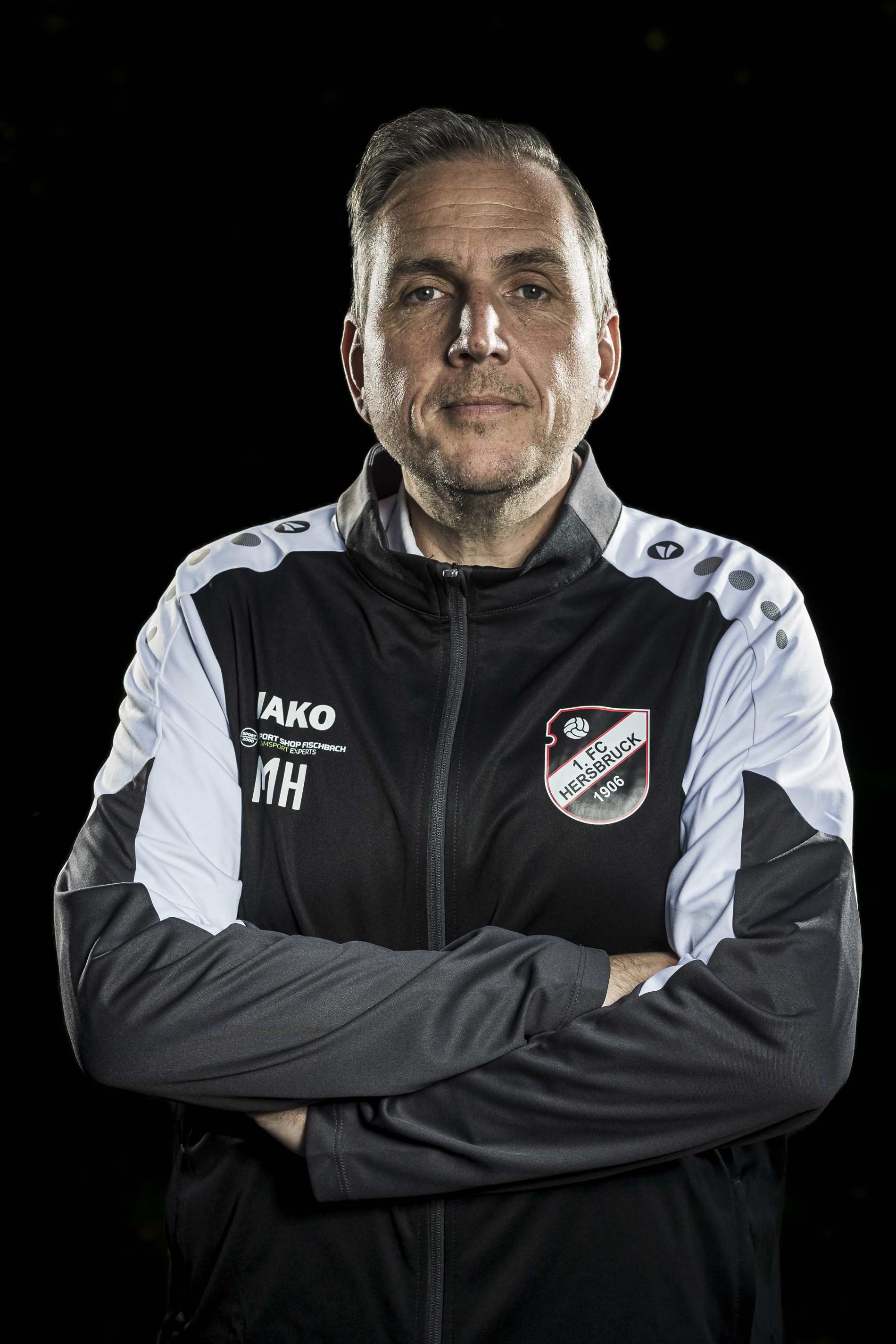Trainer des 1. FC Hersbruck 1906 in Vereinsjacke mit verschränkten Armen vor schwarzem Hintergrund