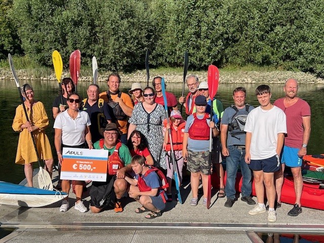 Gruppe von Menschen beim Kanufahren, einige halten Paddel und einen großen Scheck mit der Aufschrift 'APULS' und 500 Euro, im Hintergrund ein Fluss und Bäume.