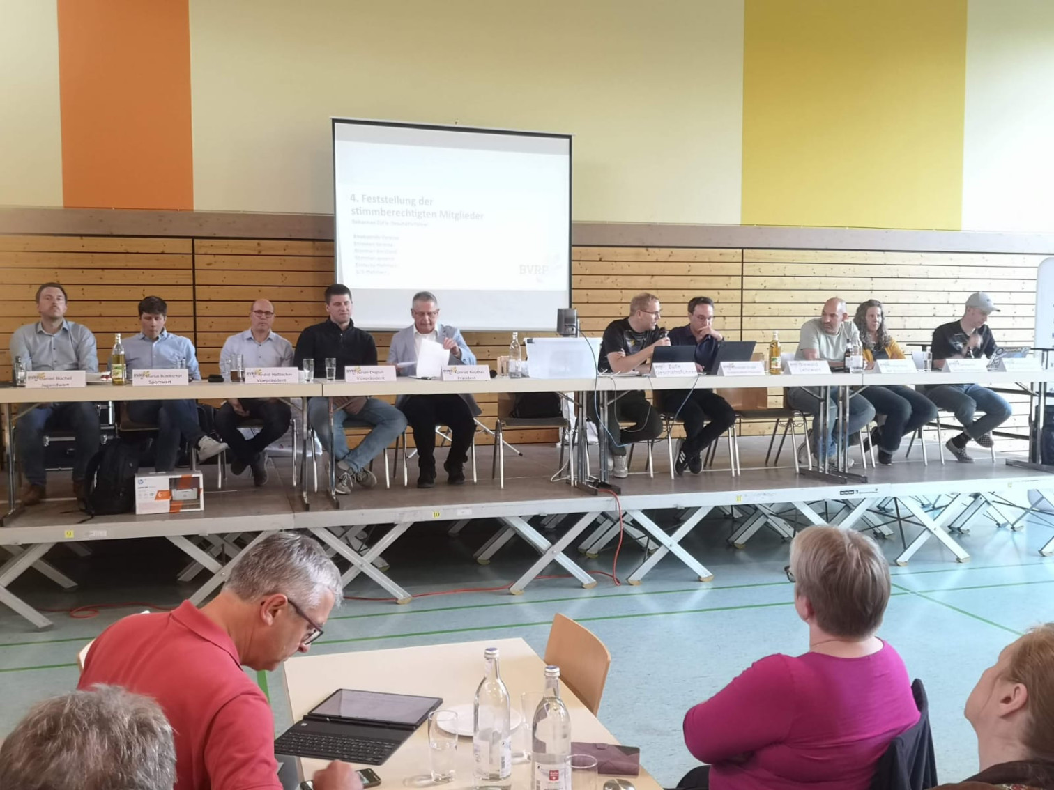 Podiumsdiskussion bei einer Vereinsversammlung in einer Turnhalle mit mehreren Teilnehmern und Präsentation auf Leinwand