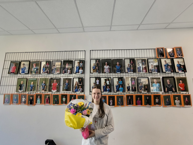 Person mit Blumenstrauß vor einer Wand mit vielen Porträtfotos in einer Schule oder Sportverein