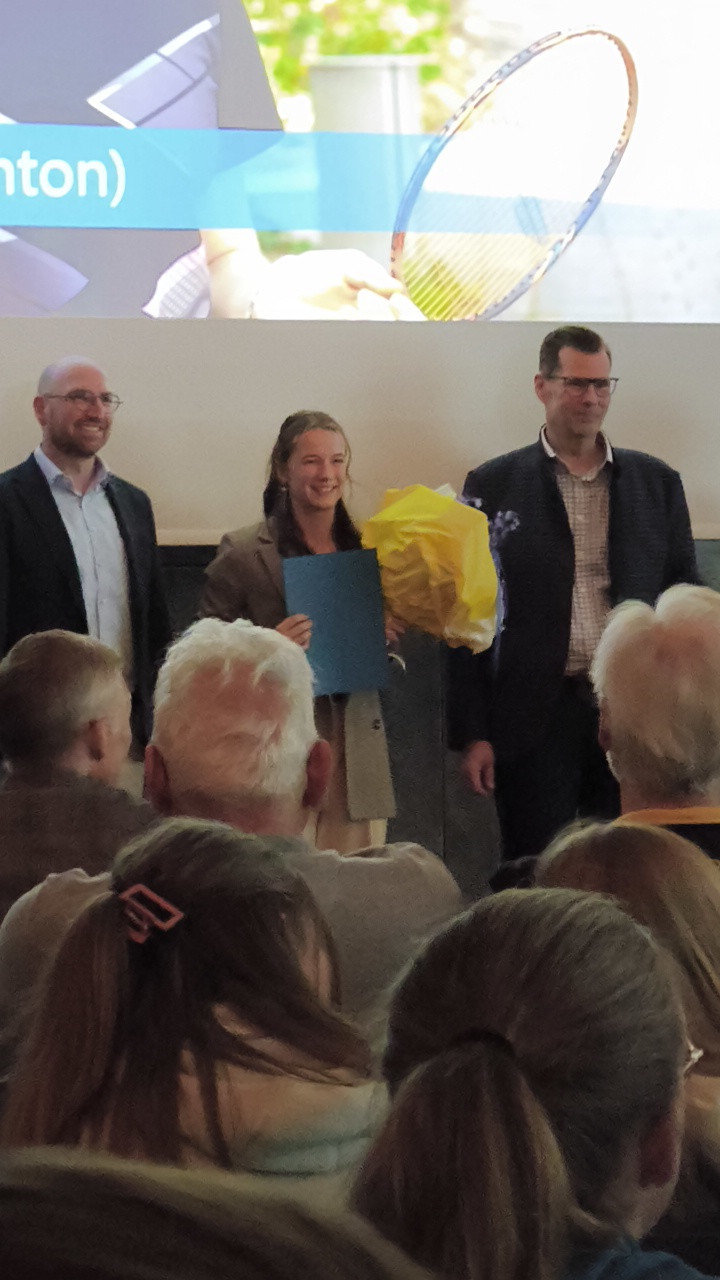 Personen bei einer Preisverleihung oder Ehrung mit Blumenstrauß und Urkunde vor Publikum