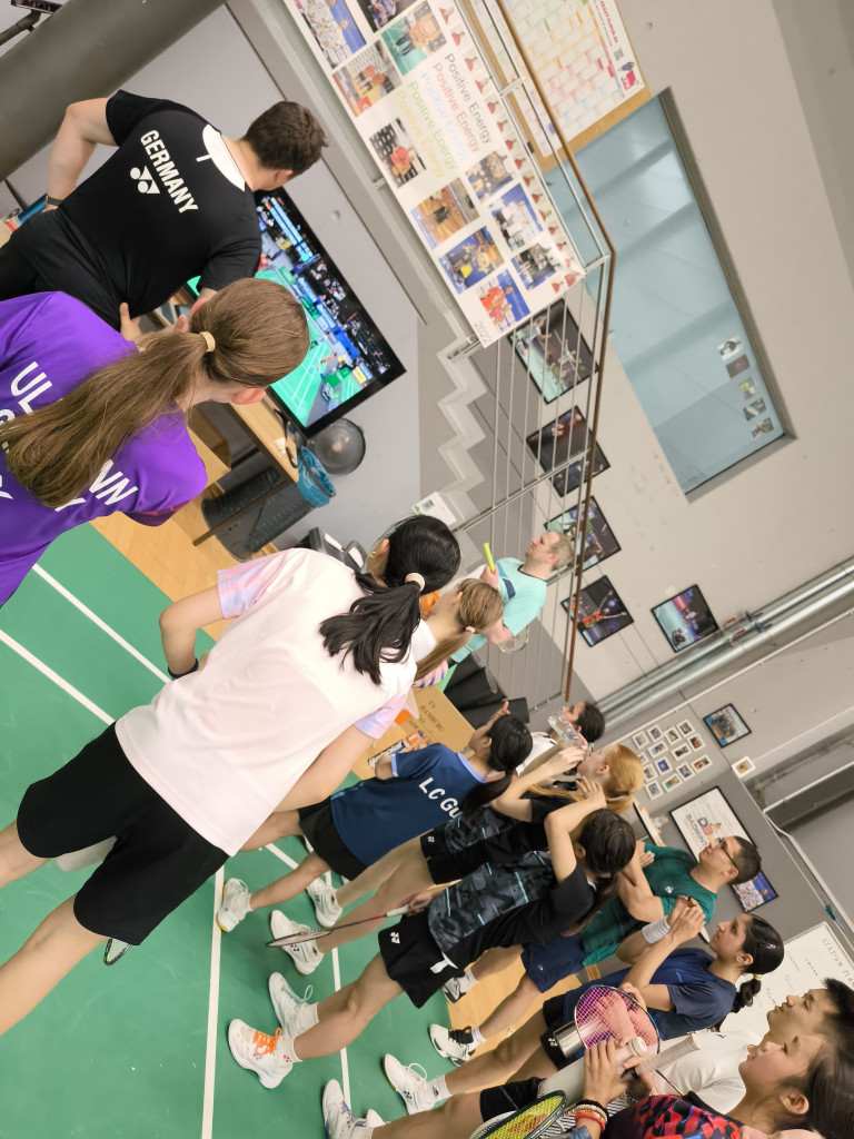 Jugendliche beim Badminton-Training in einer Sporthalle, Trainer erklärt Taktik am Bildschirm