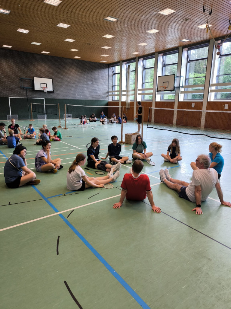 Kinder und Jugendliche sitzen mit Badmintonschlägern in einer Sporthalle im Kreis, Badminton-Training oder Schulsport