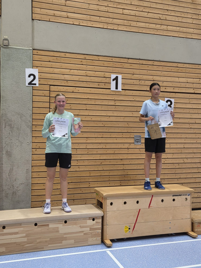 Zwei junge Sportlerinnen auf dem Siegerpodest in einer Sporthalle mit Urkunden und Preisen