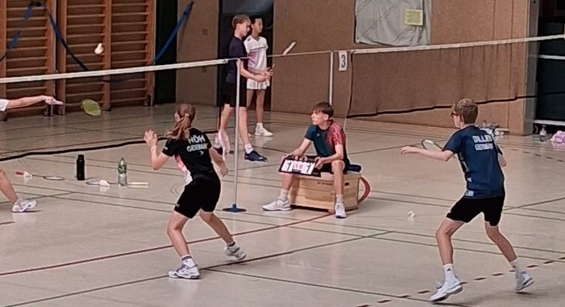Jugendliche spielen Badminton in einer Sporthalle, Schiedsrichter zählt Punkte, Spielerin mit 