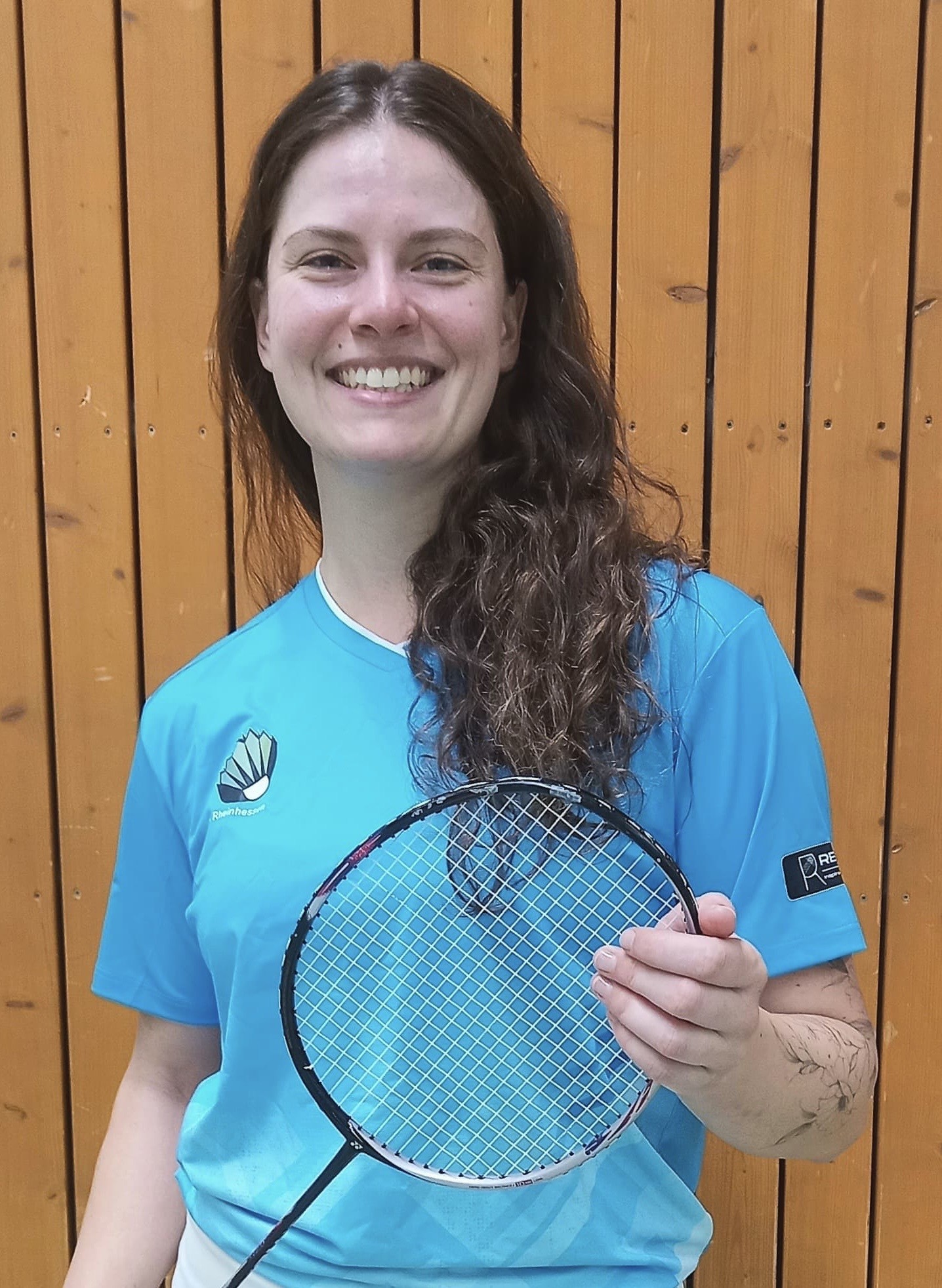 Bianca Hofert im blauen Trikot mit Badmintonschläger vor Holzpaneelen