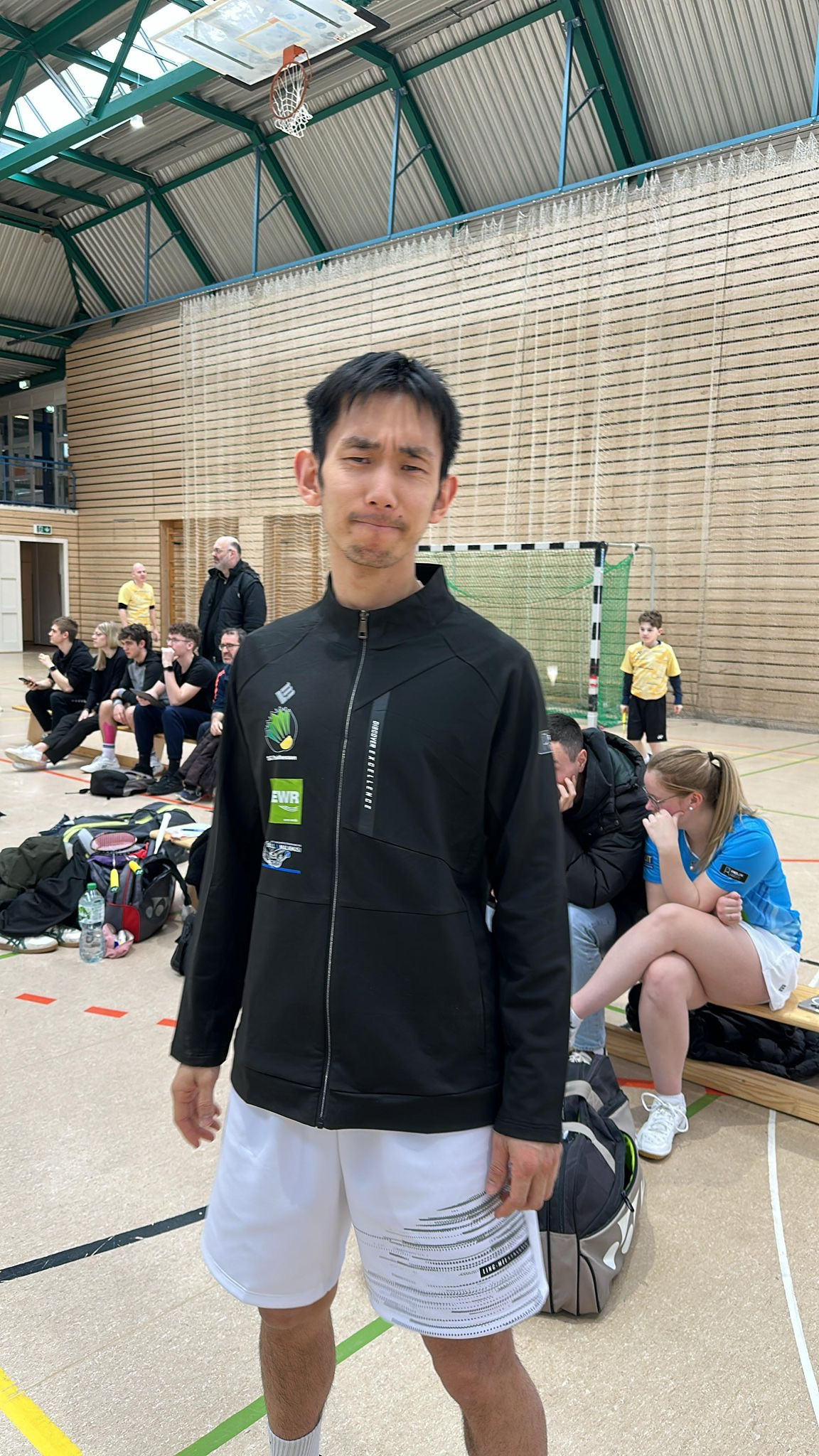 Sportler in schwarzer Jacke bei einem Turnier in einer Sporthalle mit Zuschauern und Spielern im Hintergrund