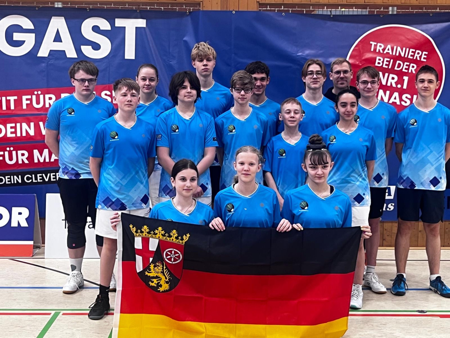 Gruppenfoto eines Sportteams mit blauen Trikots und einer Deutschlandflagge mit Rheinland-Pfalz-Wappen in einer Sporthalle