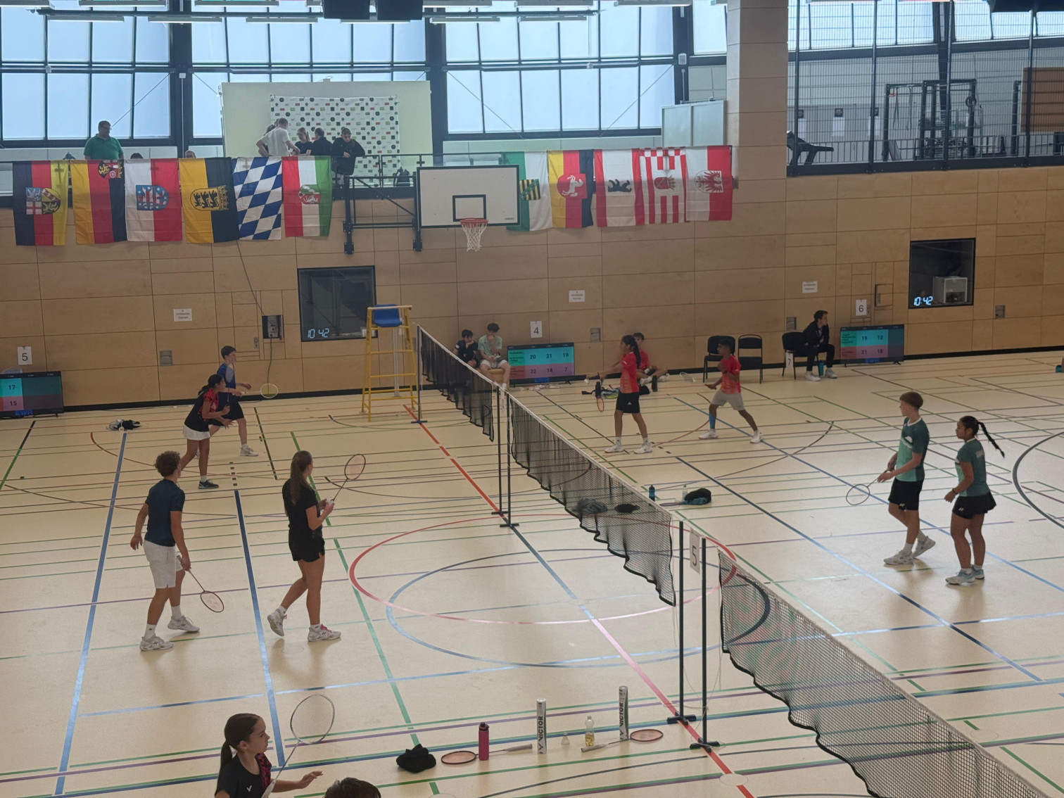 Badmintonturnier in einer Sporthalle mit mehreren Spielern und Landesflaggen an der Wand