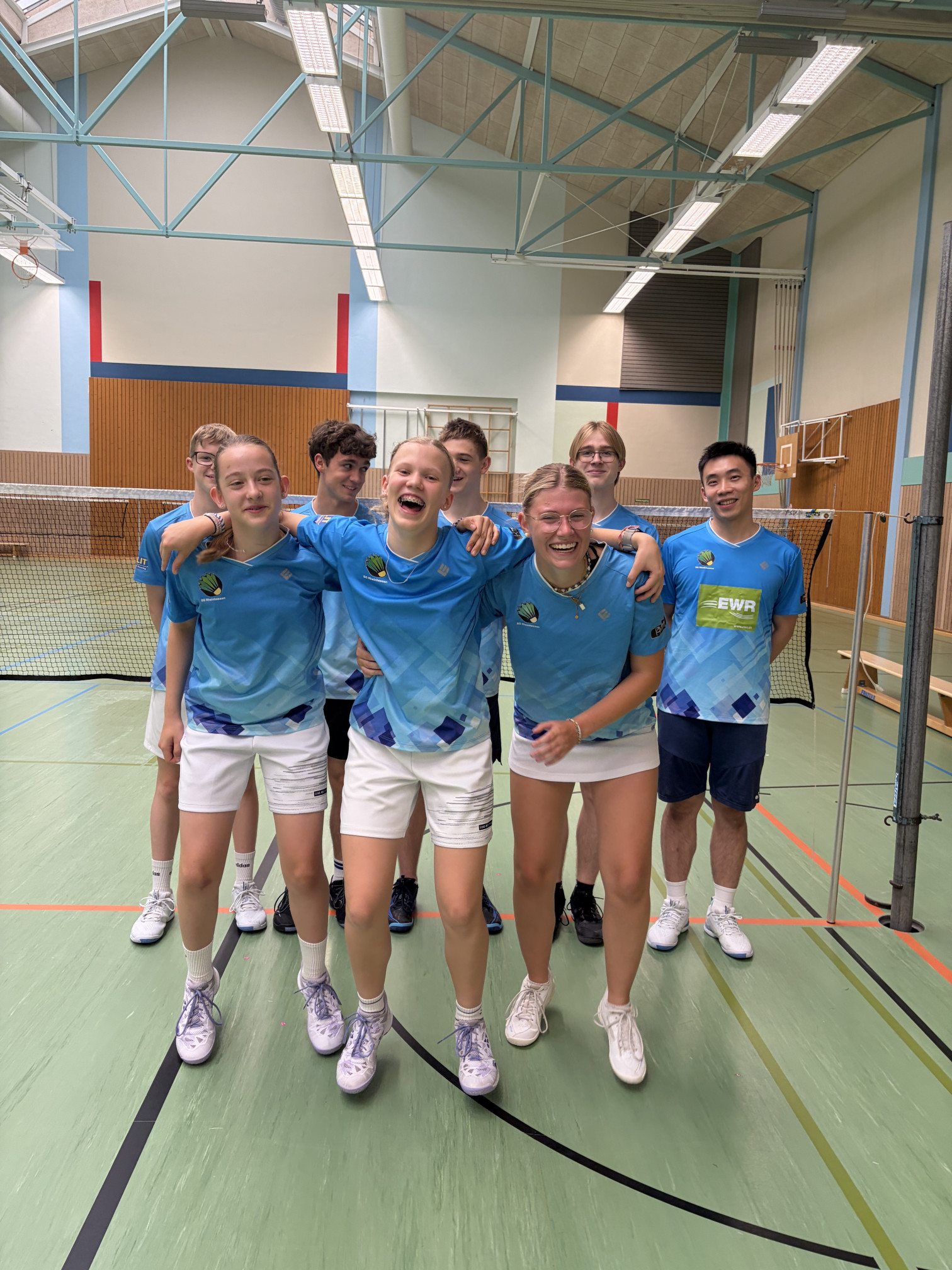 Junge Badminton-Mannschaft in blauen Trikots lachend in einer Sporthalle