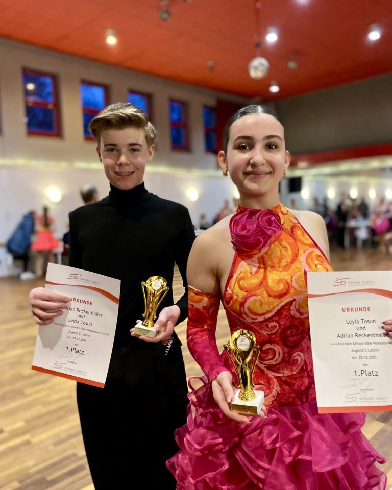 Adrian Reckenhalter und Leyla Tosun gewinnen den 1. Platz bei einem Tanzturnier und halten Urkunden sowie Pokale in den Händen.