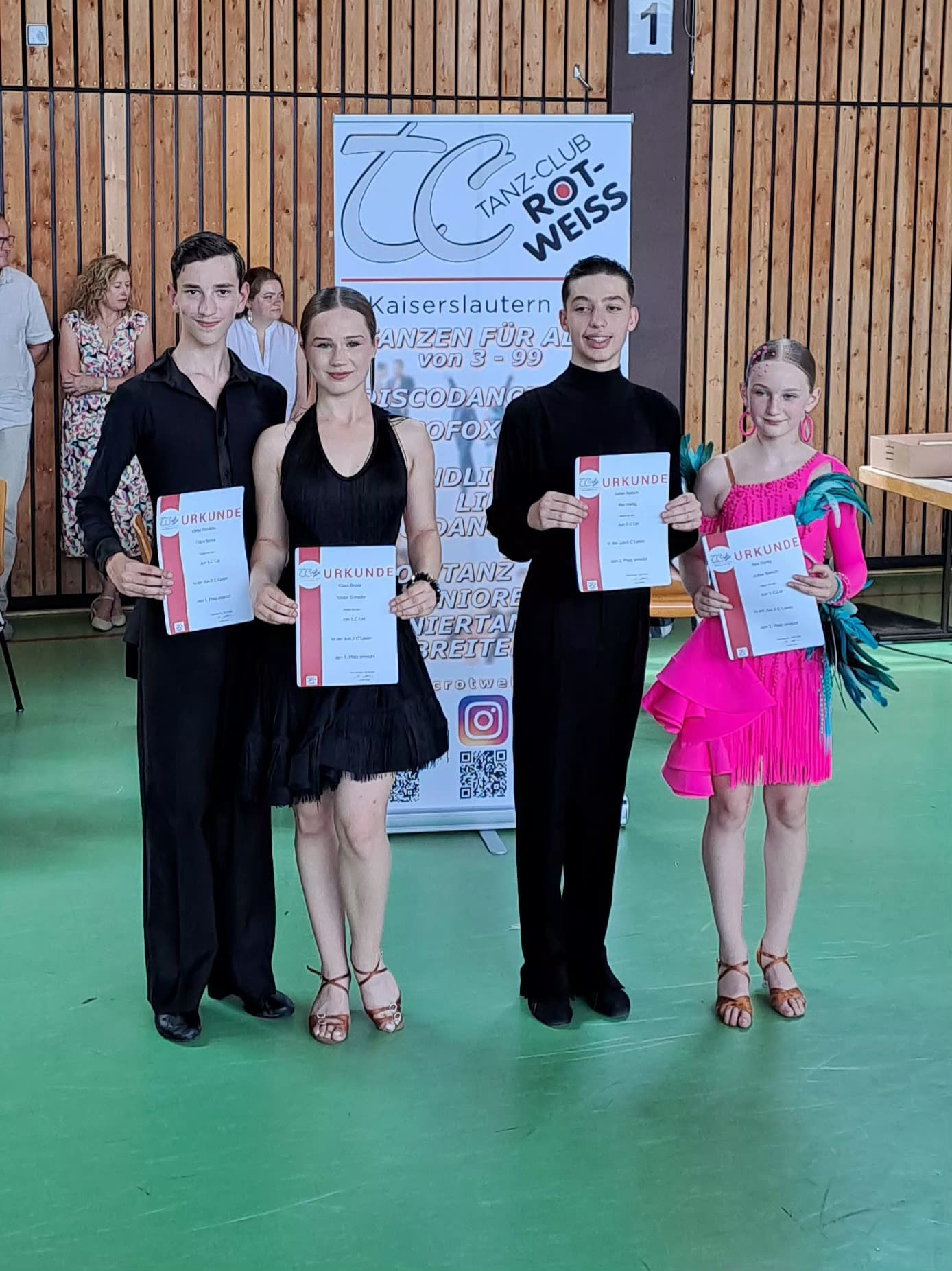Viktor, Clara und Katata bei einer Tanzsportveranstaltung in Kaiserslautern mit Urkunden vor einem Banner des Tanzclubs Rot-Weiss.