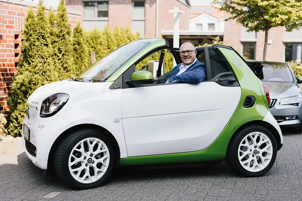 Jörg Hinrichs sitzt im Firmenwagen, einem weißen und grünen Smart Cabrio, vor einem modernen Gebäude