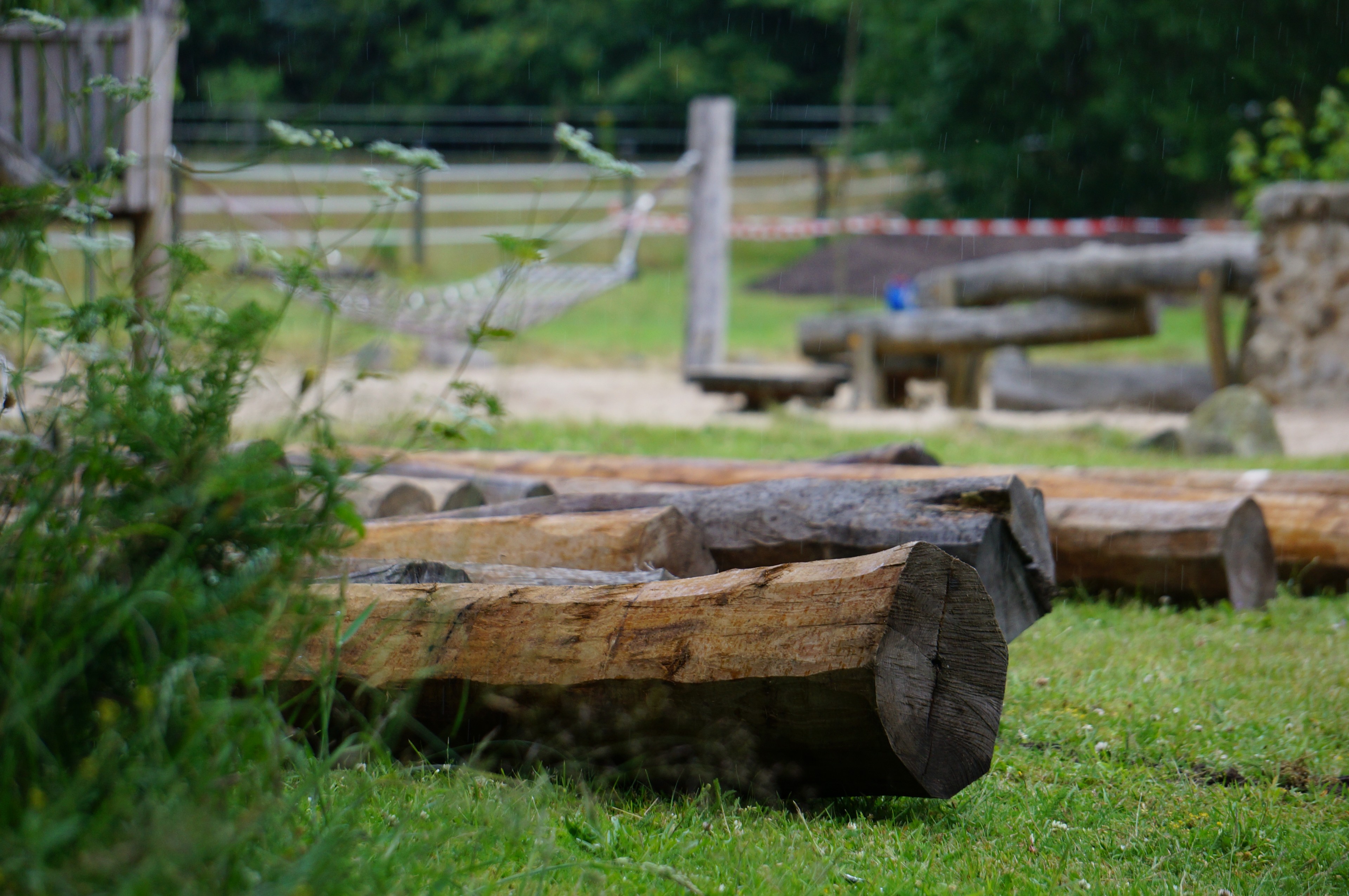 Holzstämme auf einer Wiese in einem naturnahen Spielplatz mit Hängematte im Hintergrund