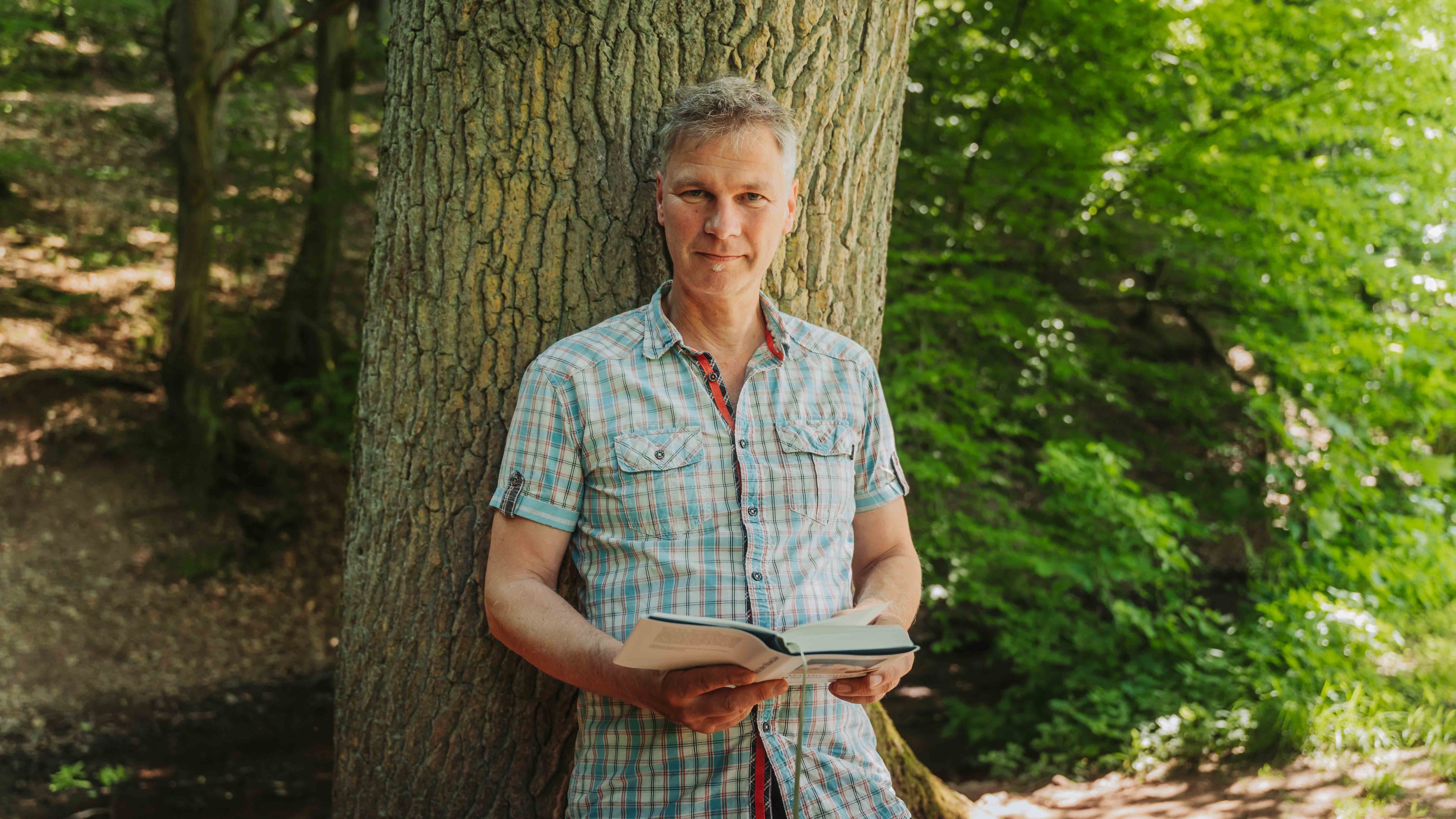 Person lehnt an Baum und liest ein Buch im Grünen – alea alternativ s 14